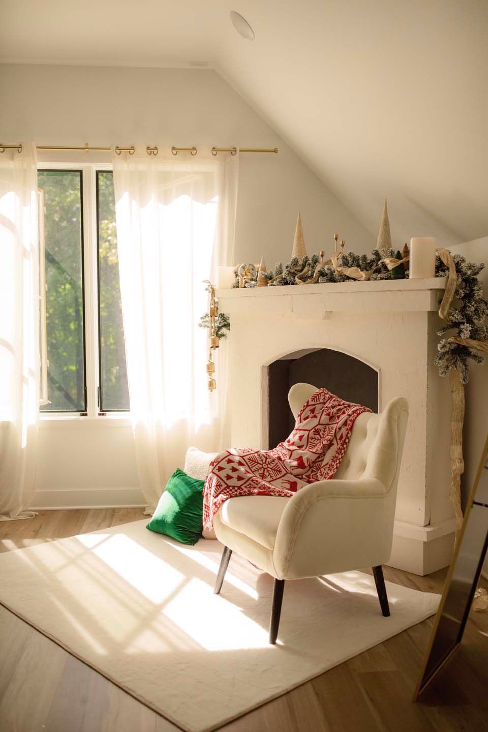 Cozy Christmas living room with white armchair, red holiday blanket, green pillow, and decorated fireplace mantel in warm sunlight.