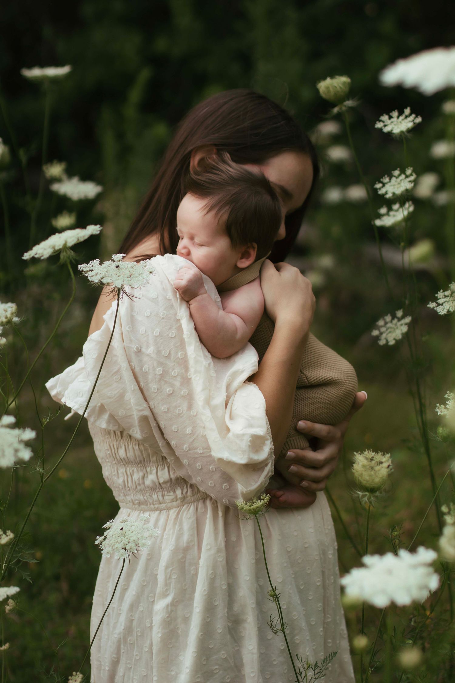 Mother in cream dress tenderly holds sleeping newborn baby among white wildflowers in lush green field.