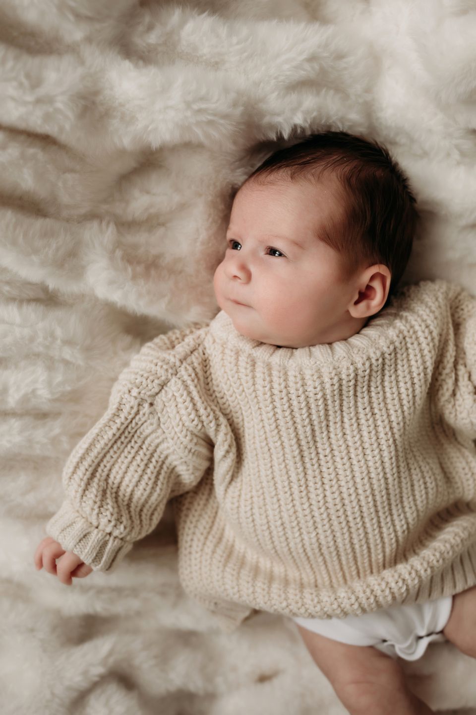 Alert infant wearing cozy cream sweater rests on soft textured white blanket.