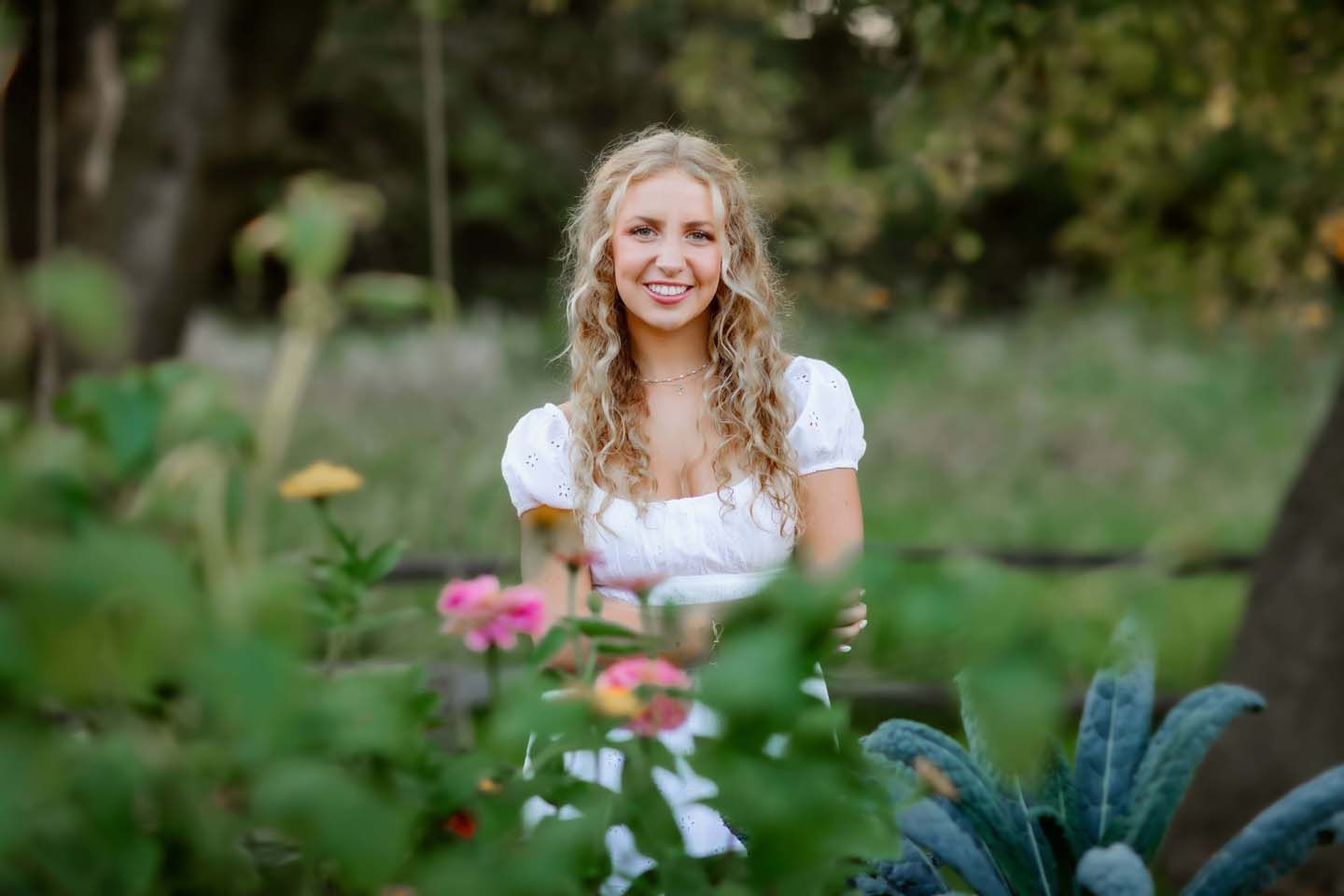 Portrait in white summer dress surrounded by blooming garden flowers and greenery.