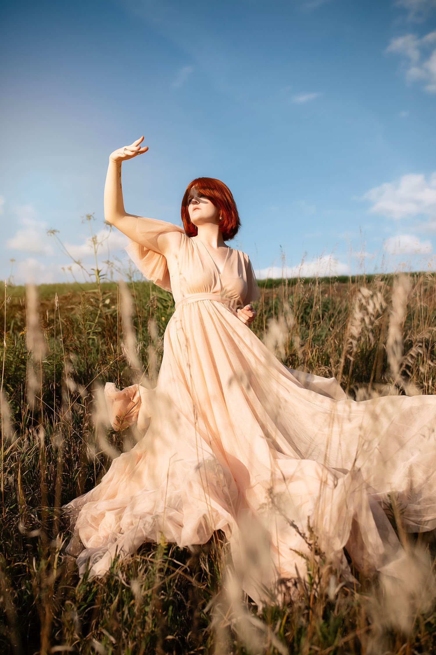 A flowing peachy-pink dress billows dramatically in a sunlit field of tall grass against a blue sky.
