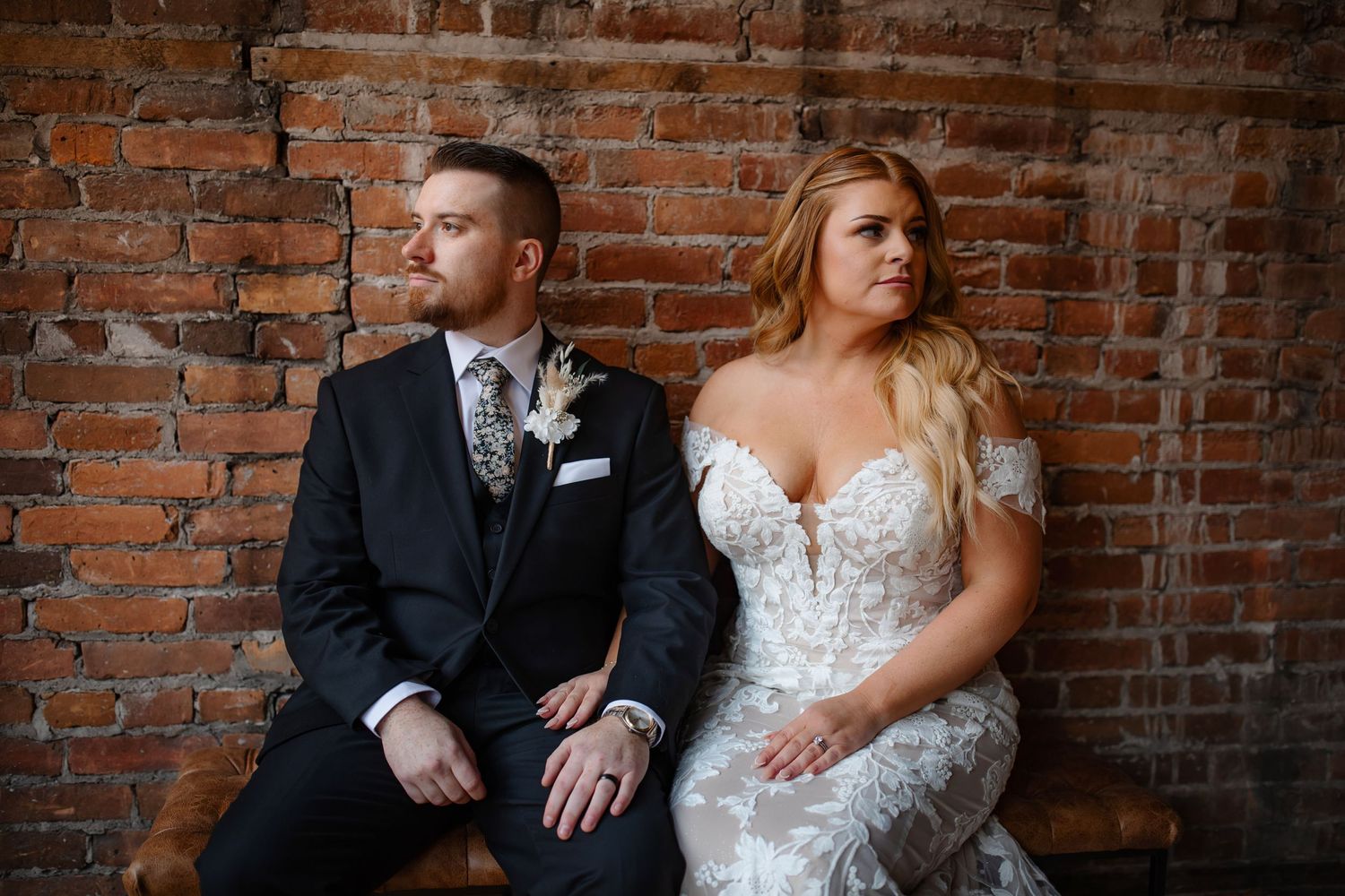 Stylish couple in wedding attire sits against exposed brick wall in urban setting.