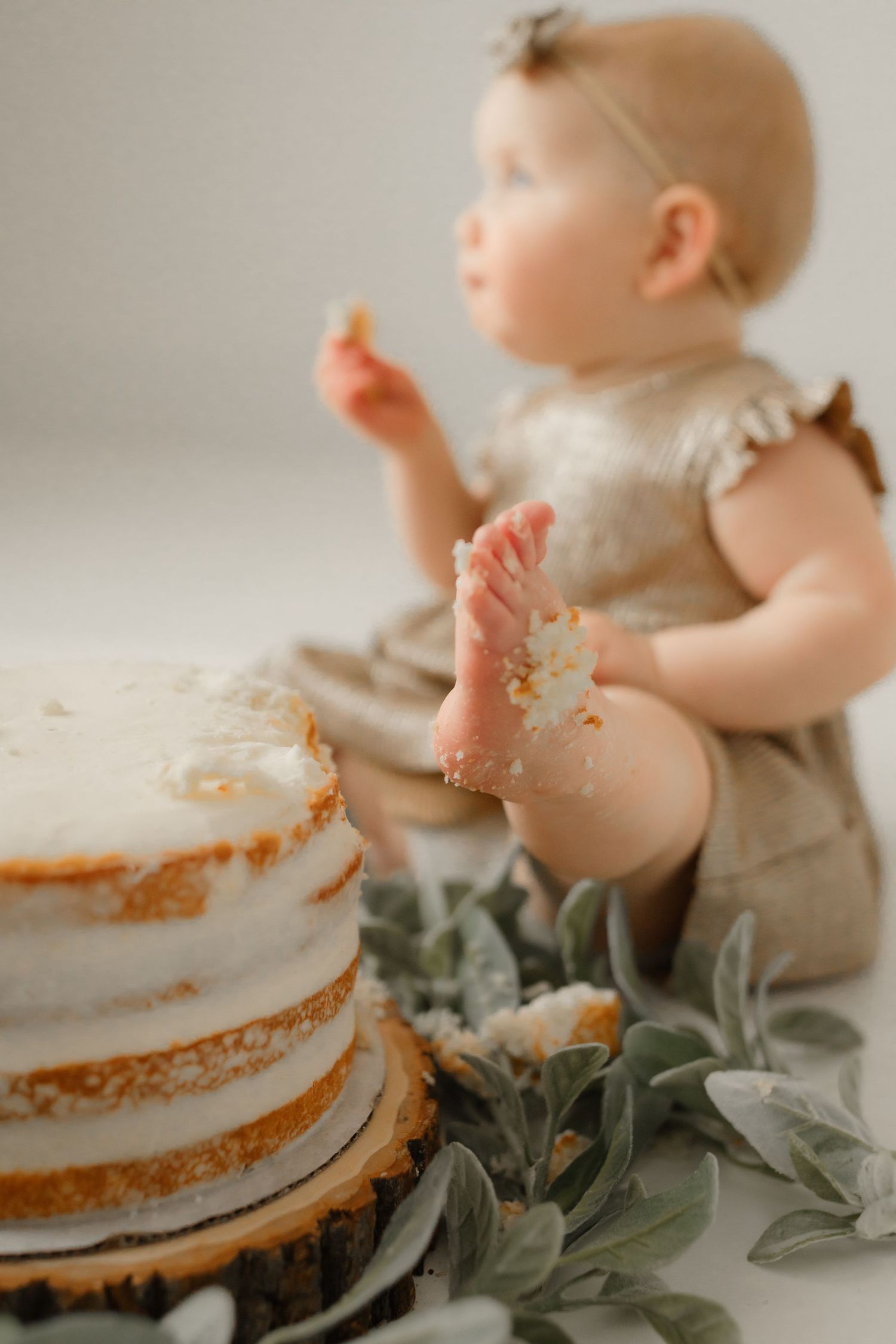 Baby next to white cake with greenery during cake smash photo session.