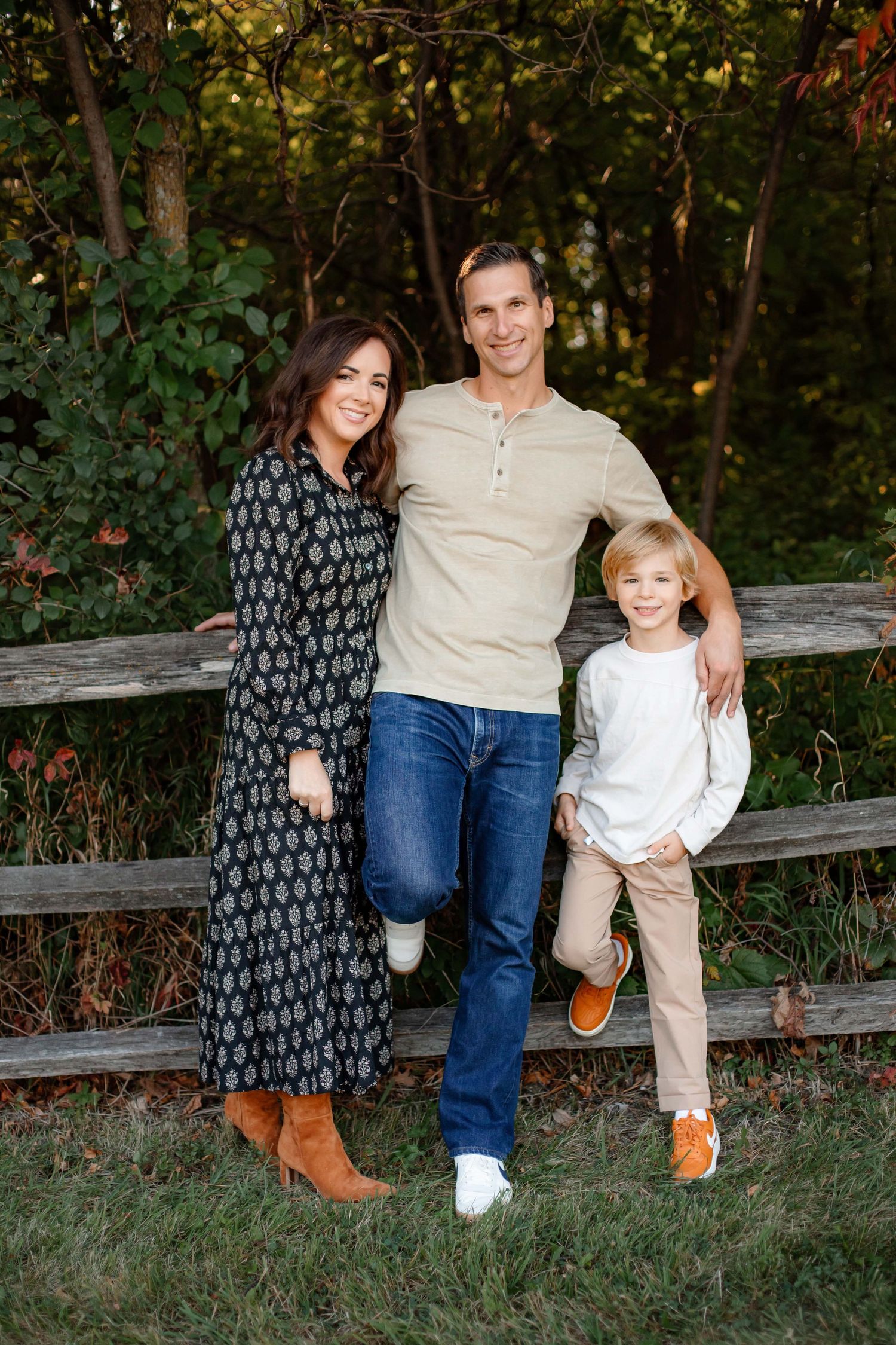 Family of three poses casually against wooden fence during outdoor photo session.