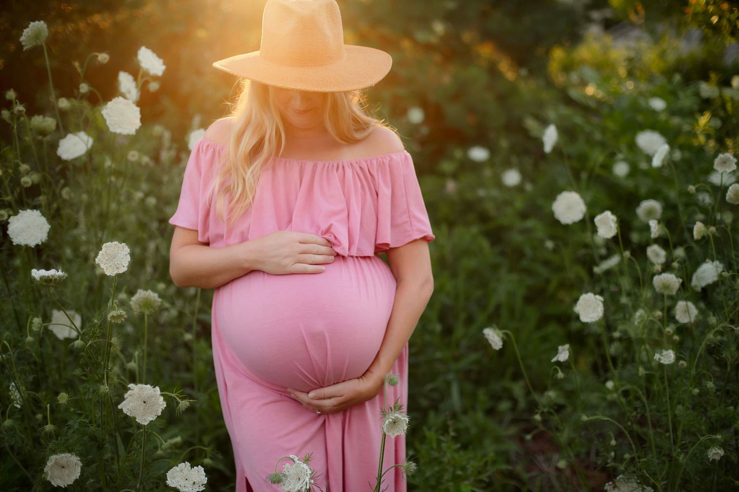 Pregnant woman in pink dress and hat holding belly in white flower field at golden hour.