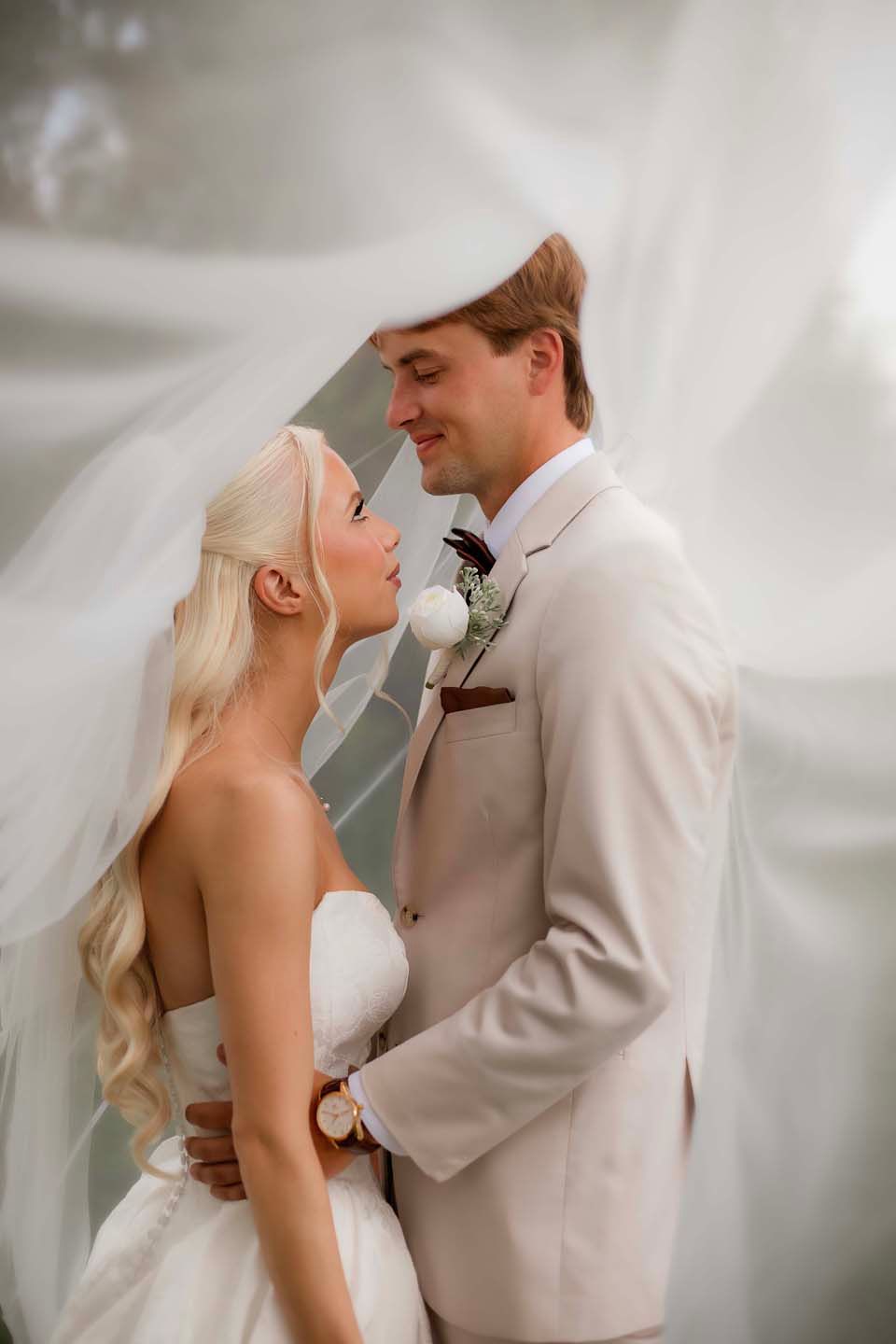 Bride and groom share romantic moment under flowing white veil, groom in beige suit with boutonniere.