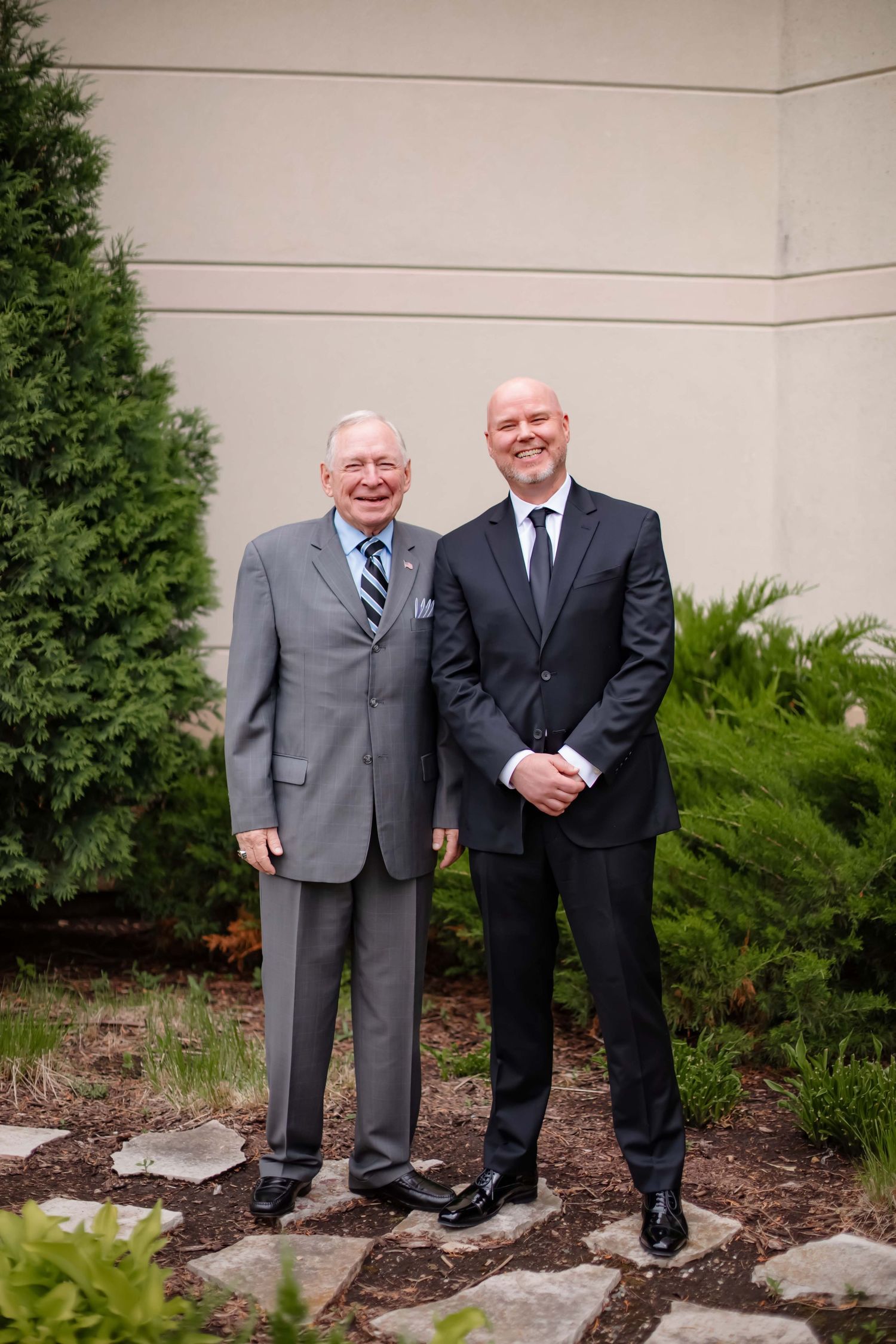 Two men in suits share a laugh while standing in front of landscaping and a white wall.