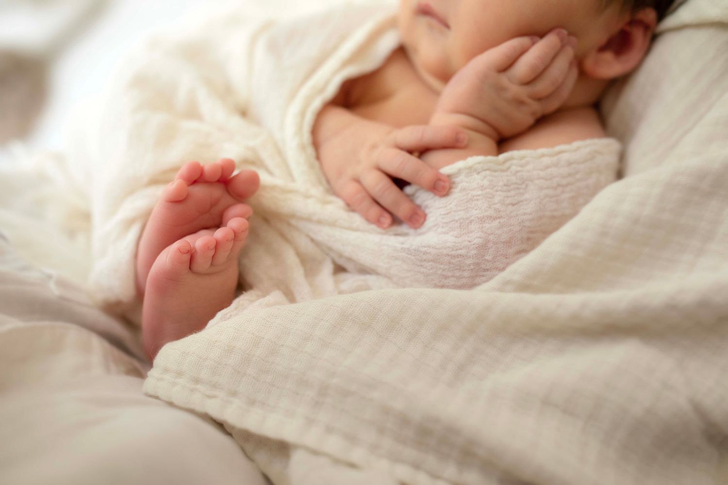 Peaceful newborn baby wrapped in soft white muslin blanket, showing tiny feet and hands while sleeping.