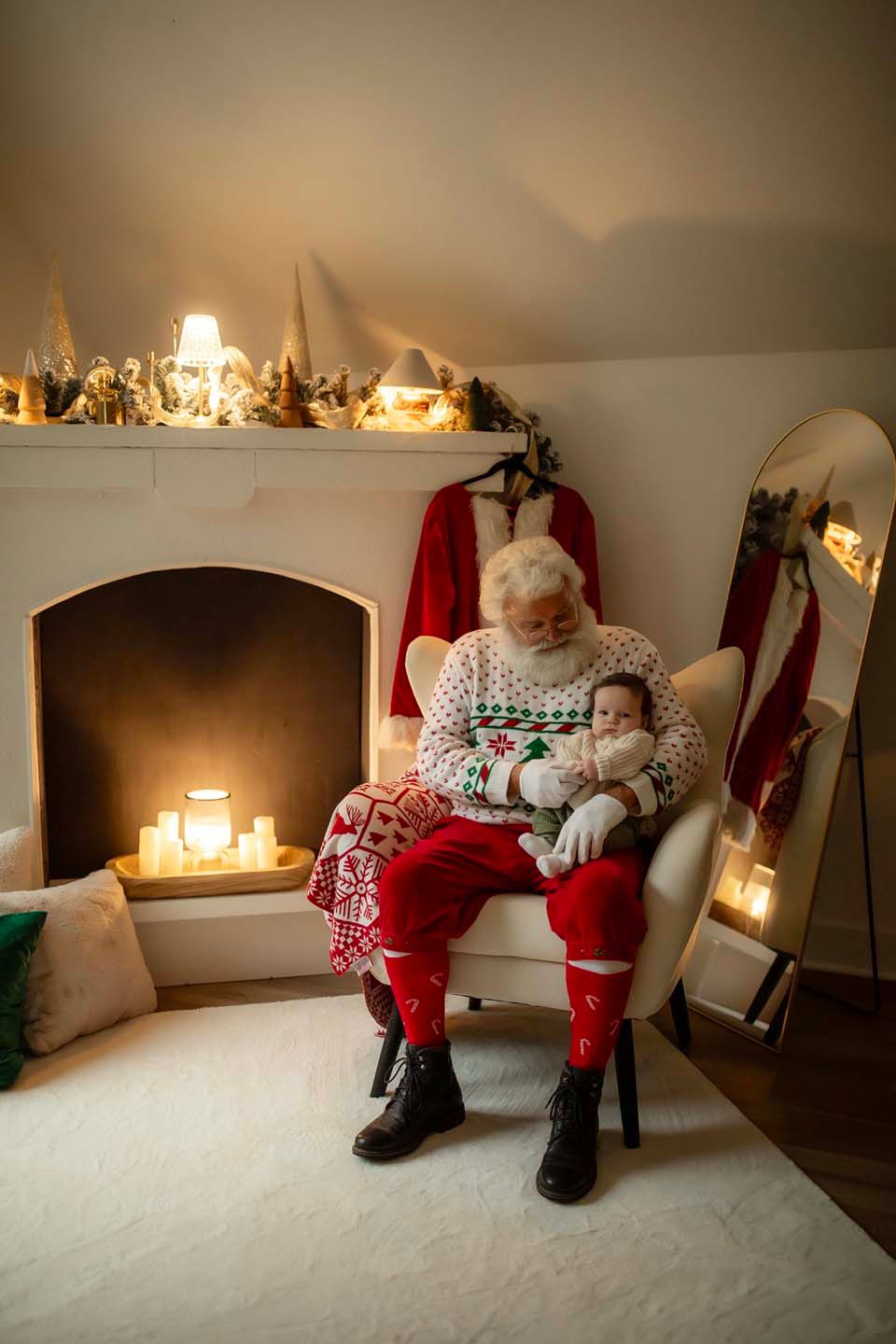 Santa Claus in festive sweater holds sleeping baby by glowing fireplace with candles and Christmas decorations.