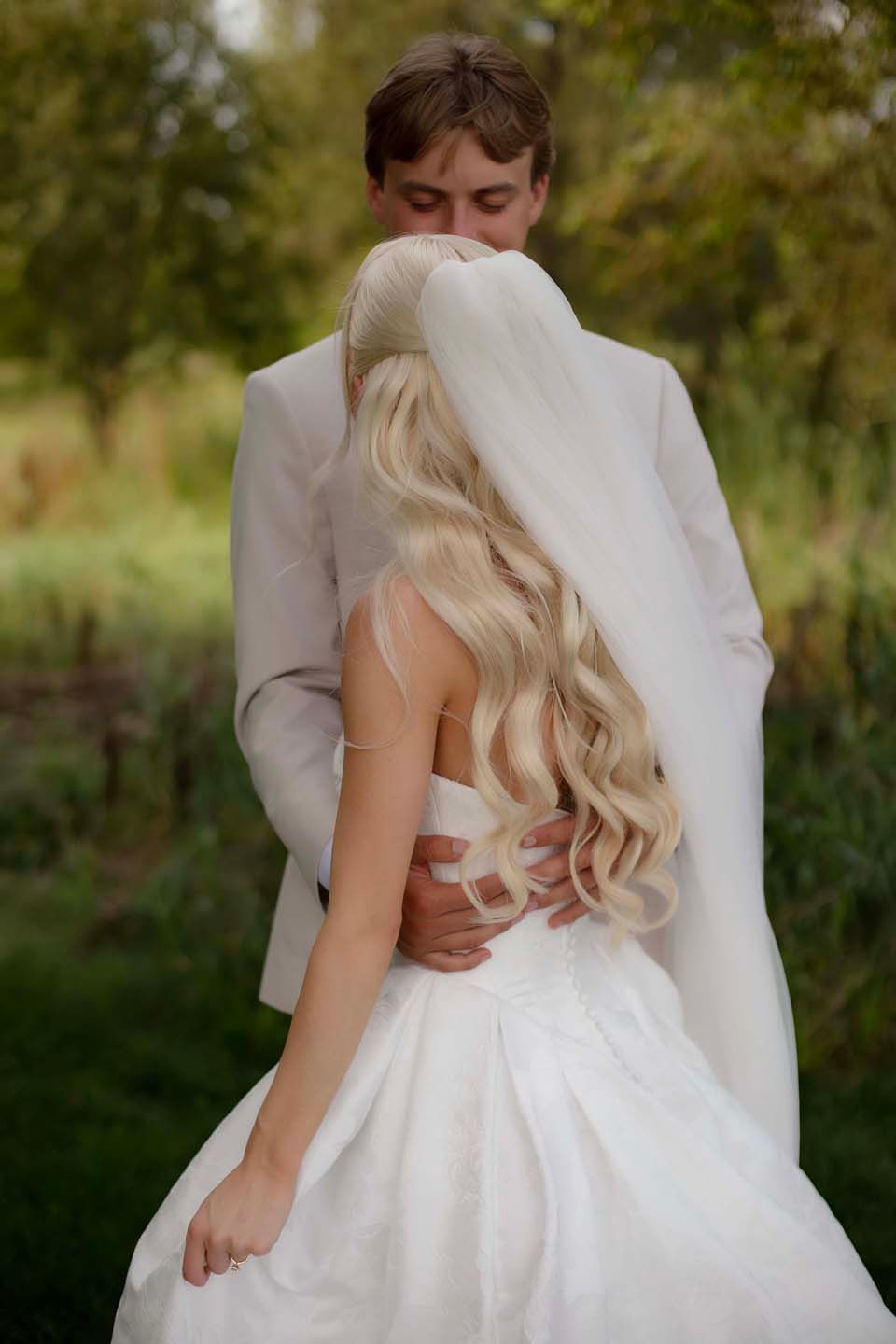 A groom in a white suit embraces his blonde bride in a flowing wedding dress and veil outdoors.