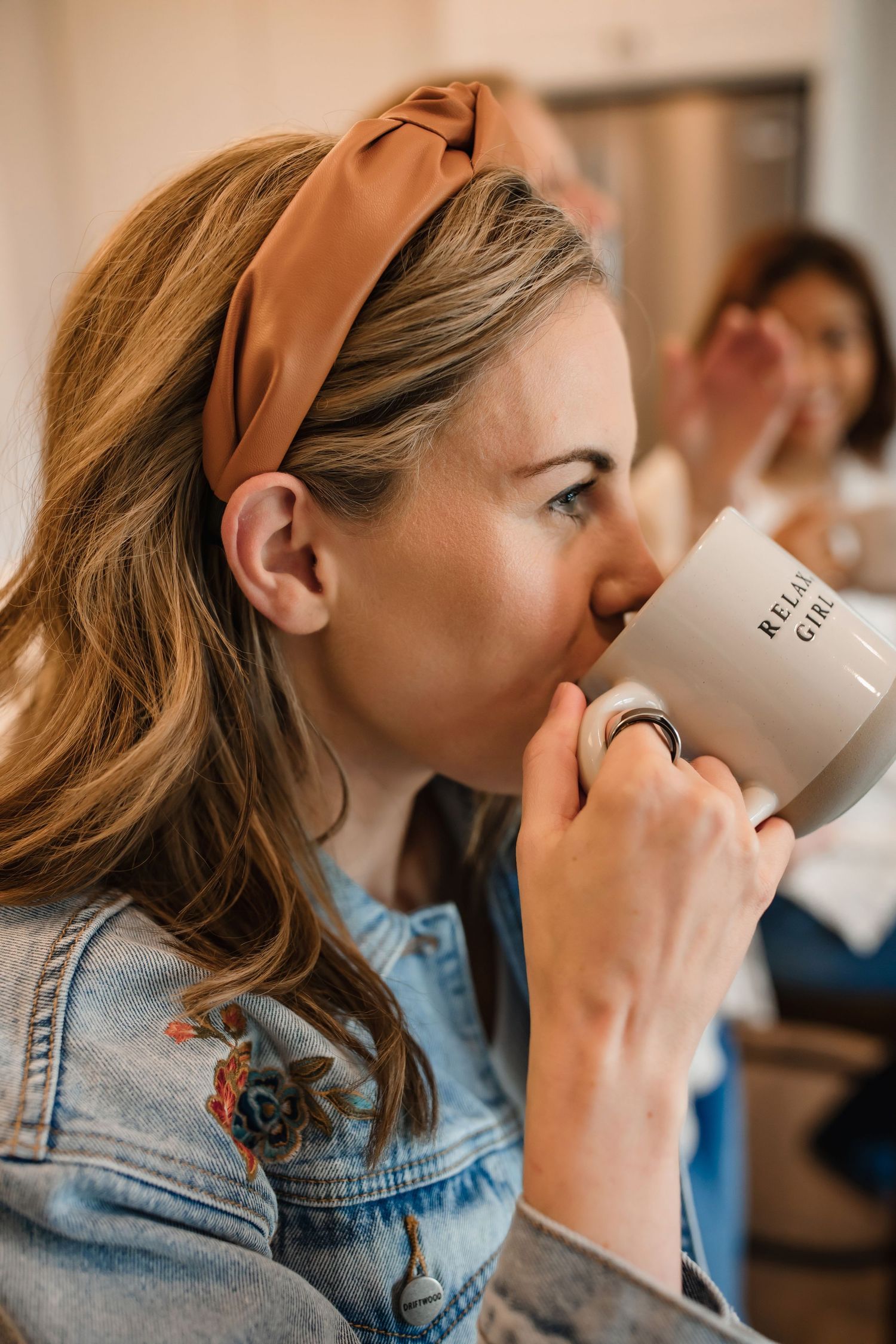 Woman in denim jacket with orange headband drinking from white ceramic mug.