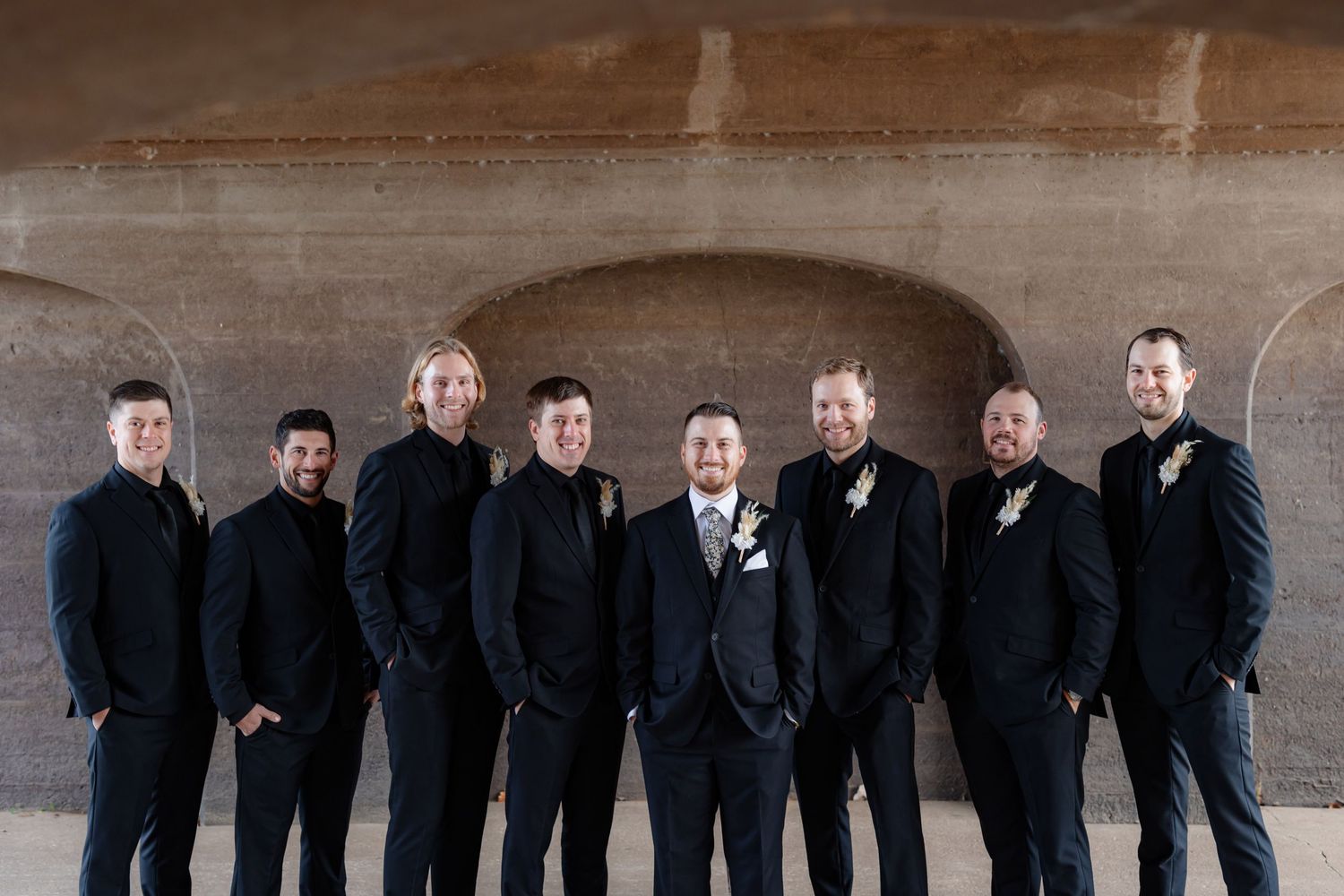 Groomsmen in black suits pose together under concrete archway.