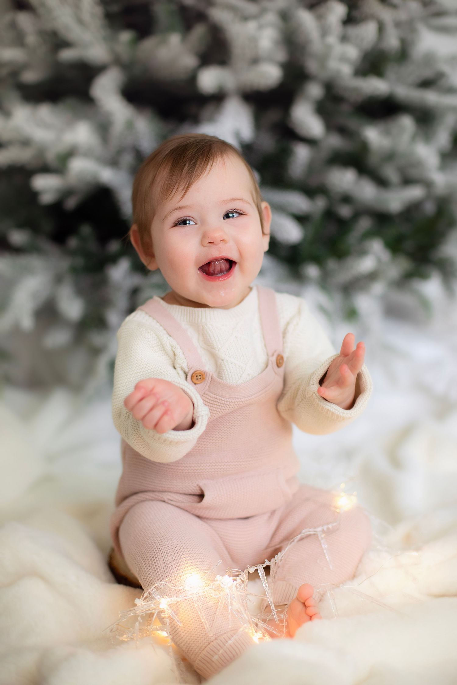 Baby in pink overalls sits on white blanket with fairy lights near snowy Christmas tree backdrop.