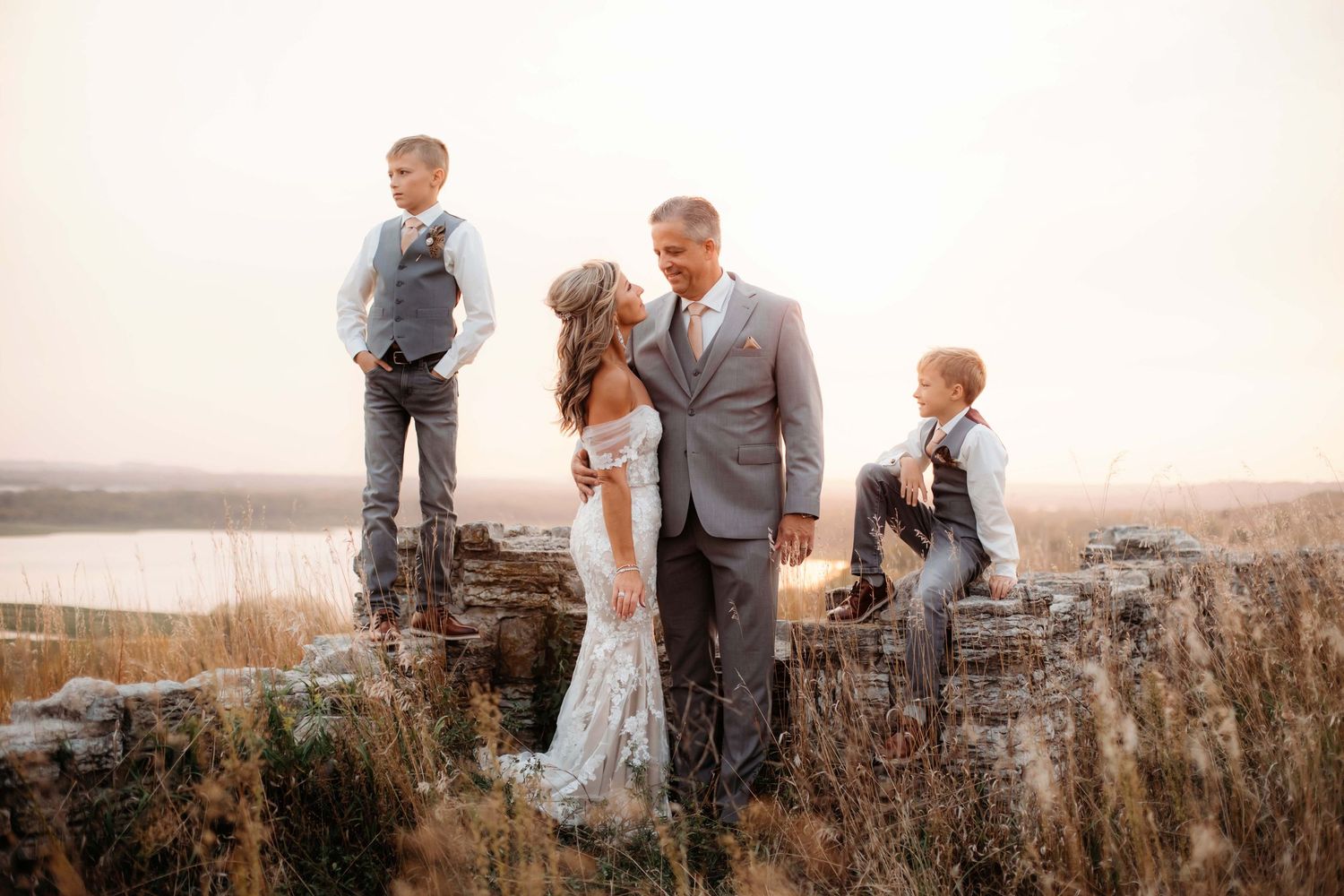 Wedding couple and children pose on stone ledge overlooking scenic landscape at sunset.