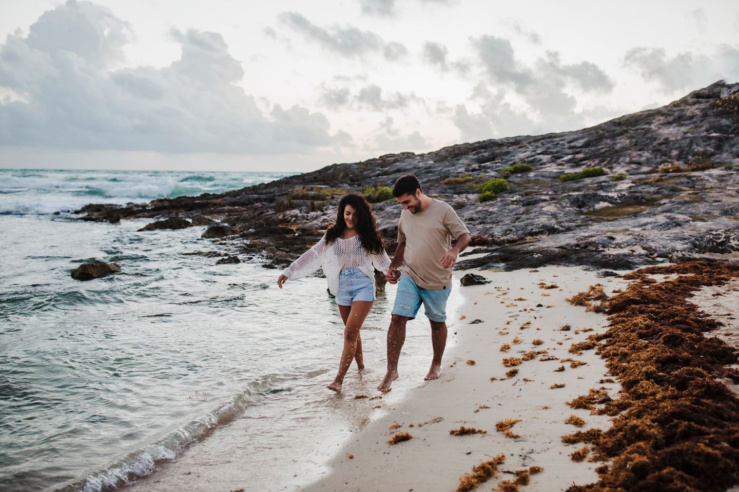 Couple running along beach shoreline with waves and rocks in background.