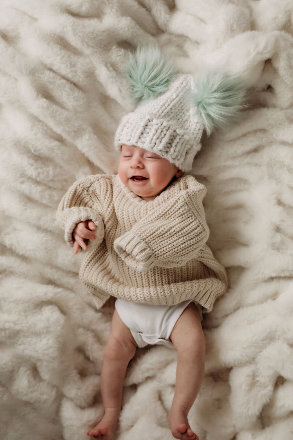 Smiling baby in cream sweater and mint pom pom hat on fluffy white blanket.