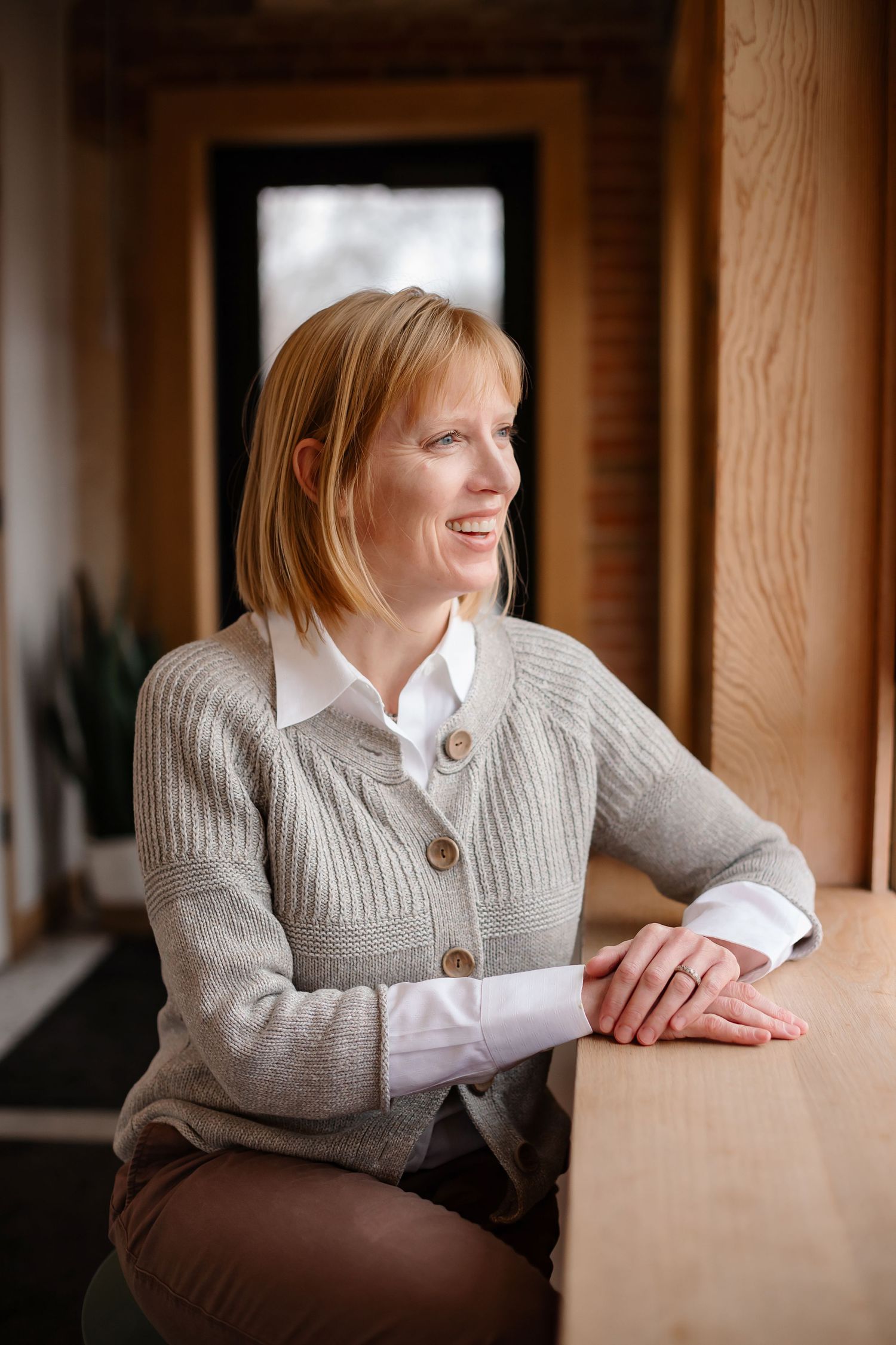 Professional wearing a gray knit cardigan and white shirt poses thoughtfully by a window.