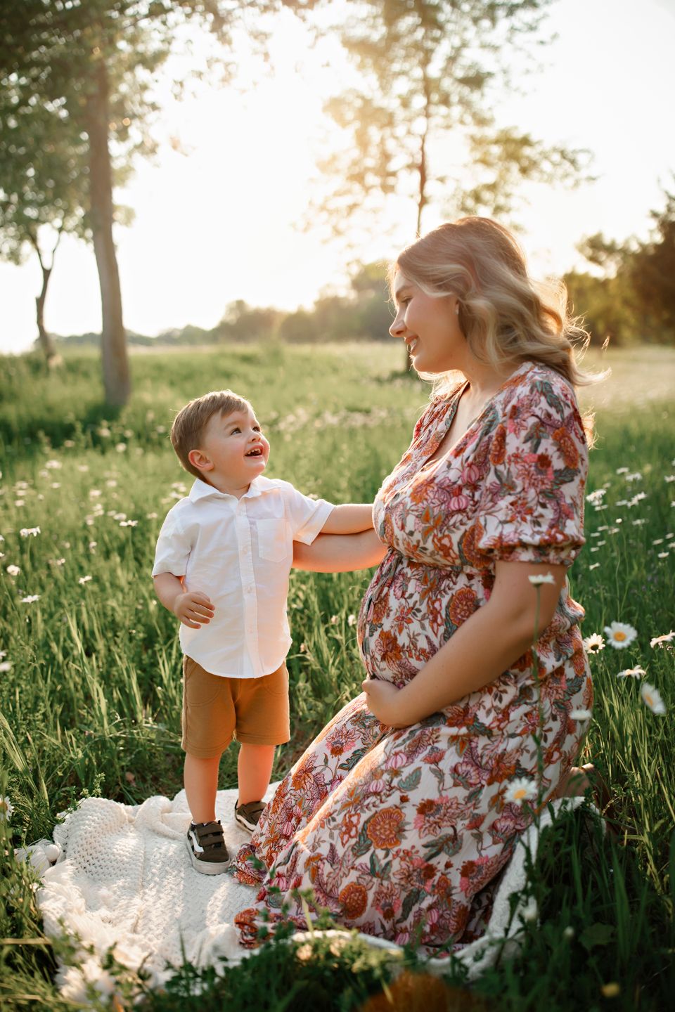 A floral dress and warm sunlight create a tender outdoor moment between parent and child in a wildflower meadow at sunset.
