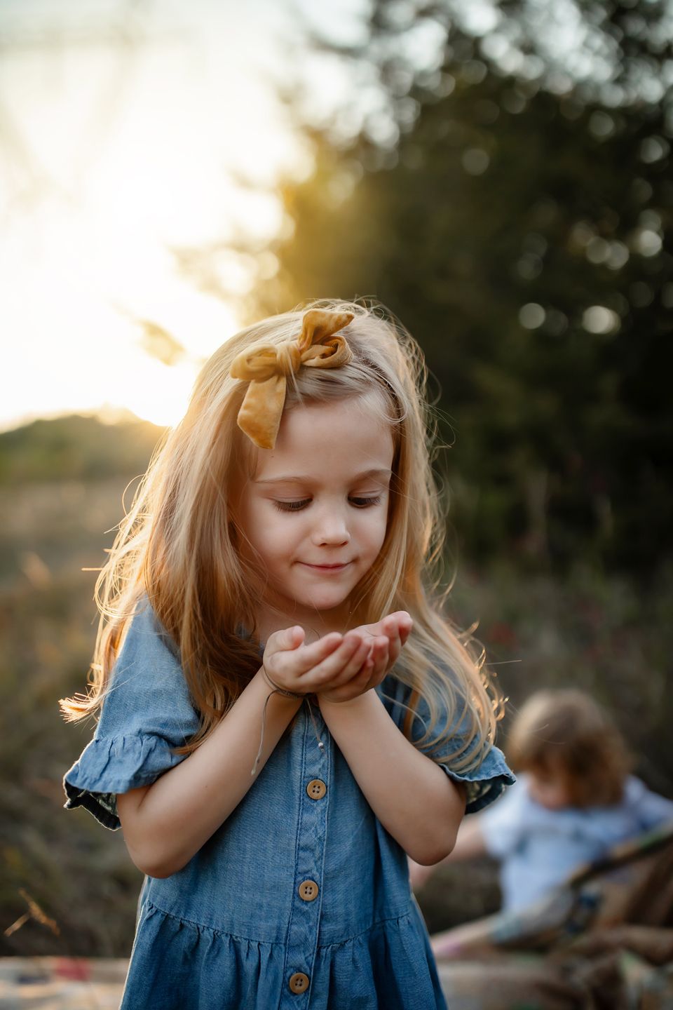 Child in denim dress holding something precious in cupped hands during golden hour in nature setting.