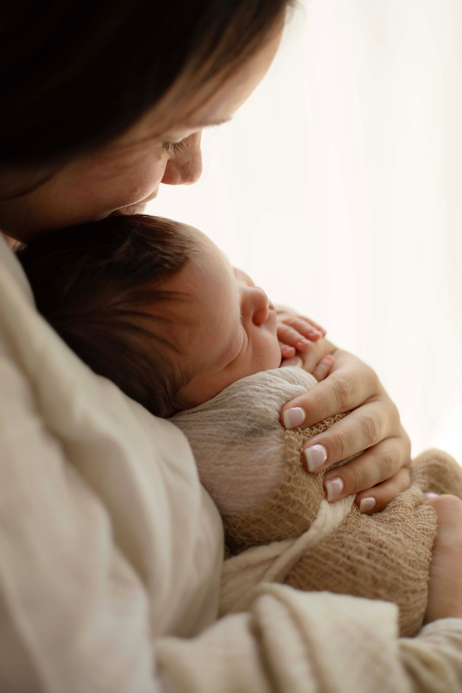 A mother tenderly holds her sleeping newborn baby wrapped in a soft beige knit blanket near a bright window.