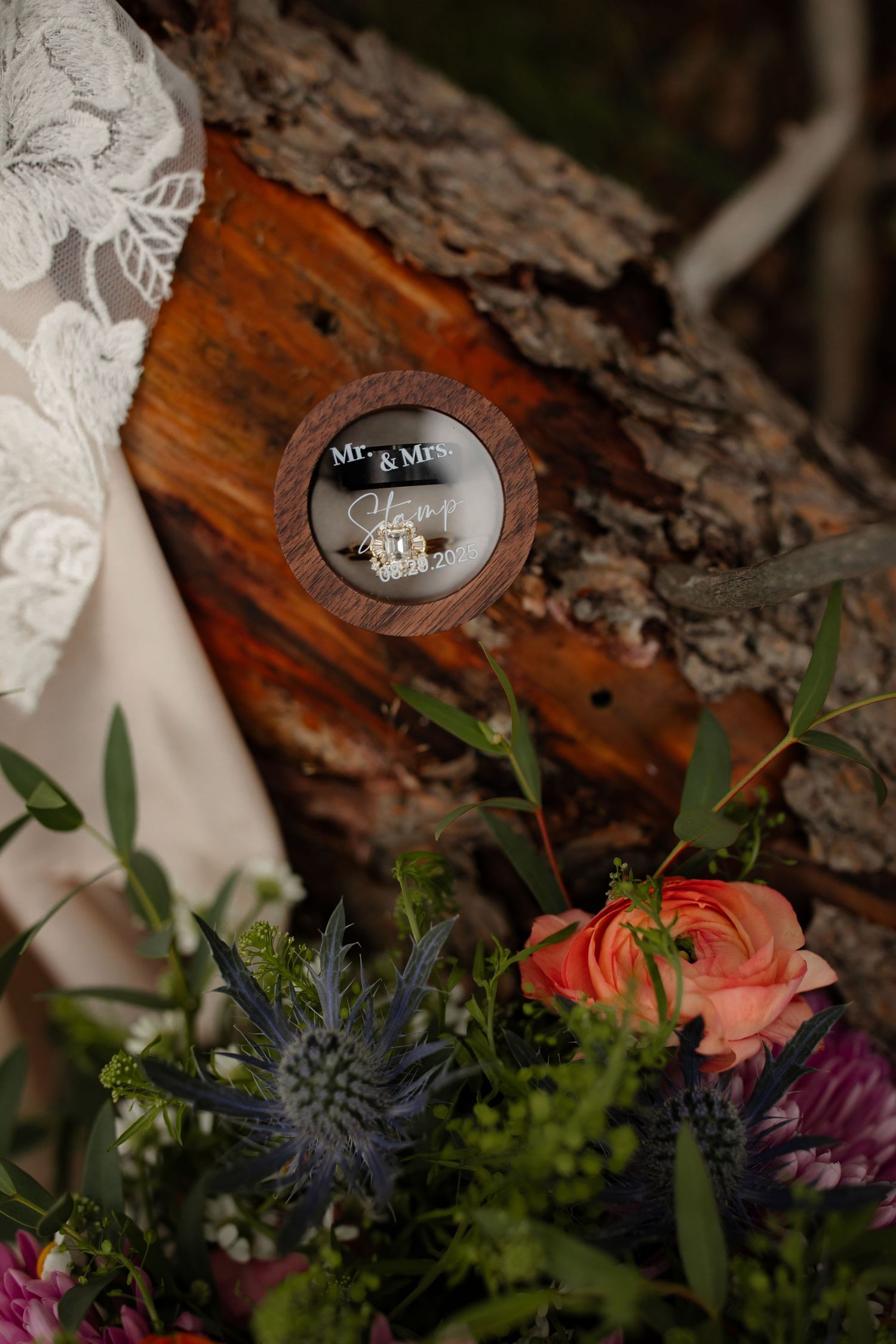 Wedding rings displayed on rustic wood with coral roses, purple flowers and blue thistle floral arrangement.