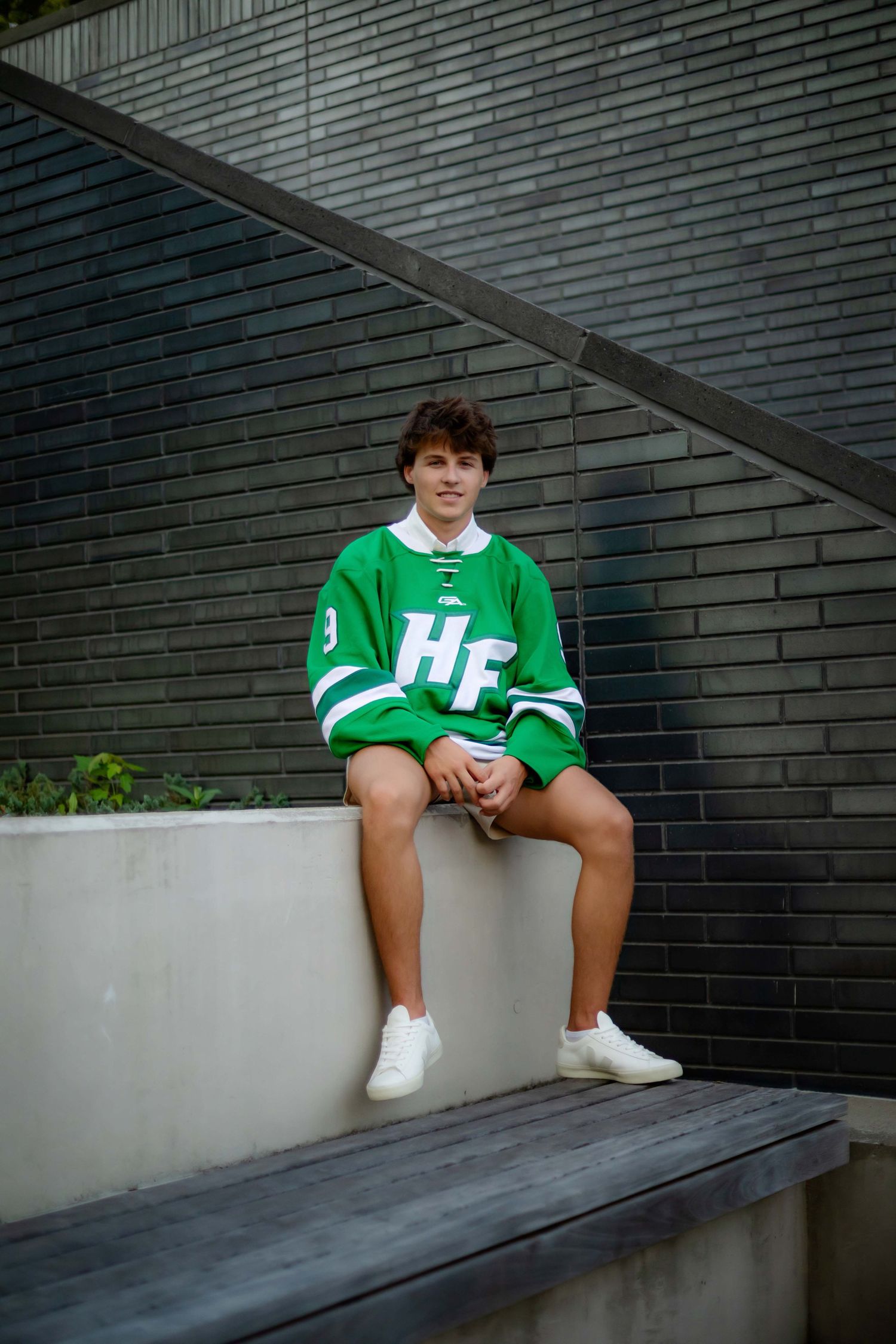 Young man in green HF hockey jersey sitting on a concrete ledge outdoors near a dark brick wall.