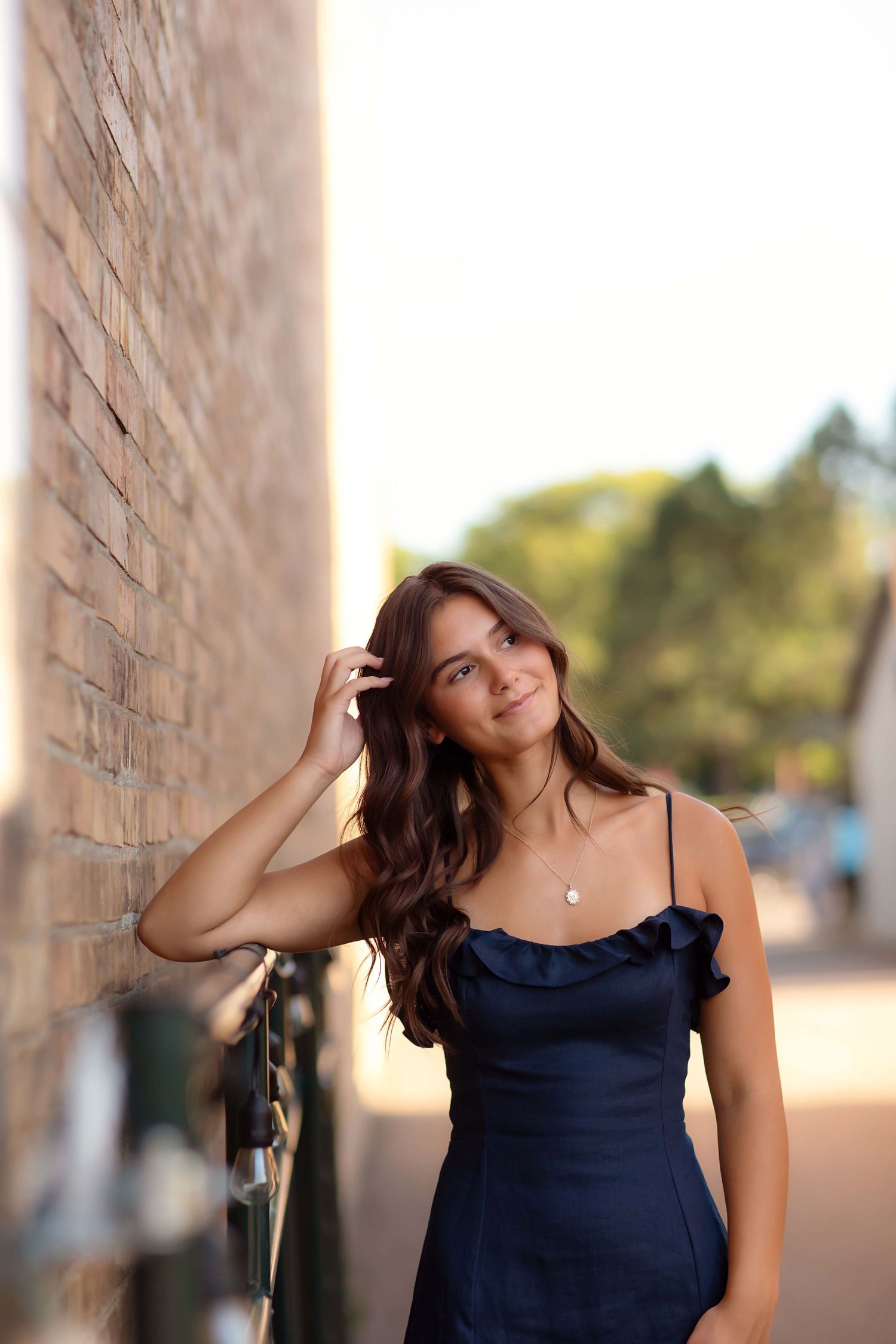 Young woman in navy ruffle dress leaning against brick wall, smiling outdoors with bokeh background.