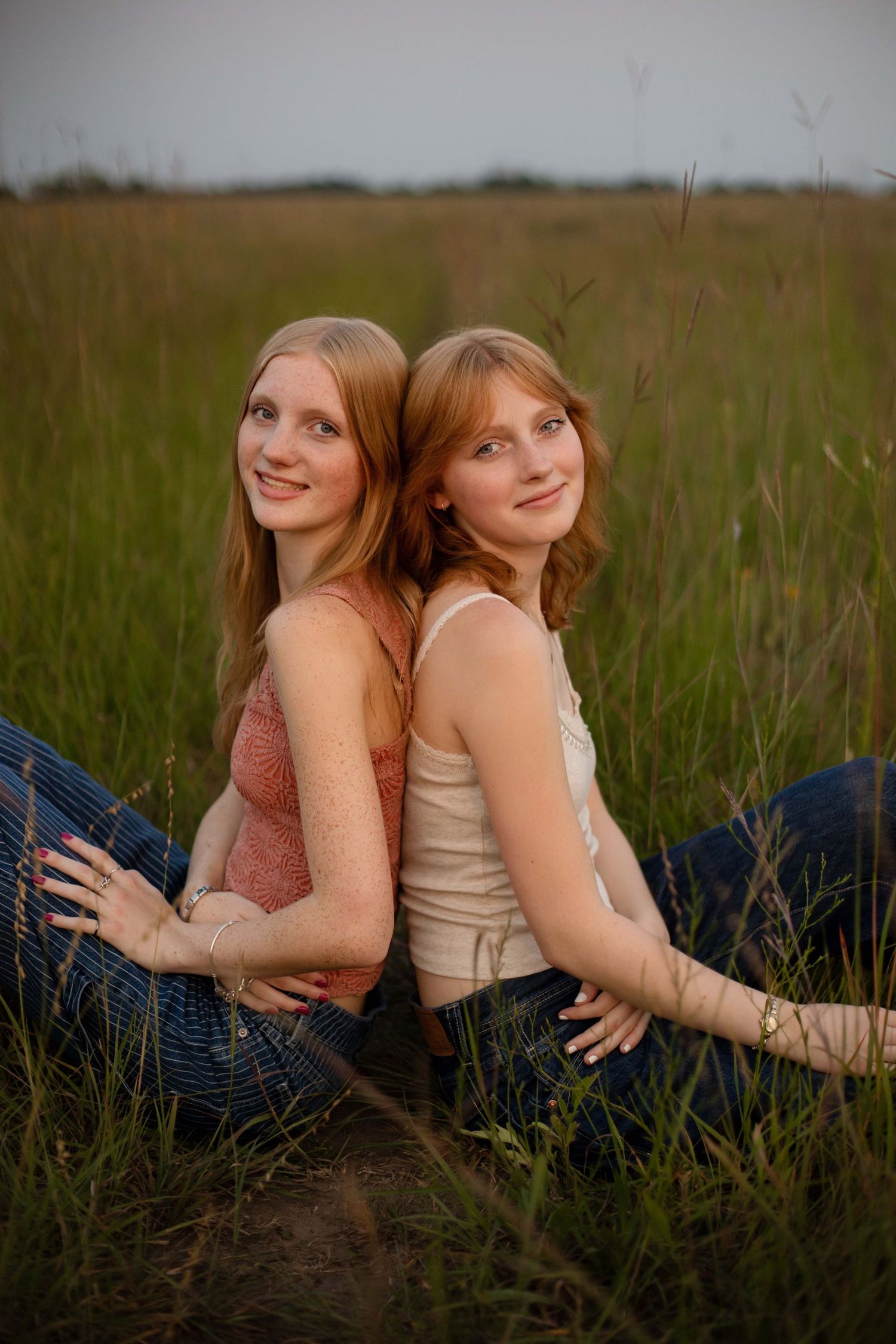 Two young women with blonde and red hair sitting back-to-back smiling in a lush green grass field.