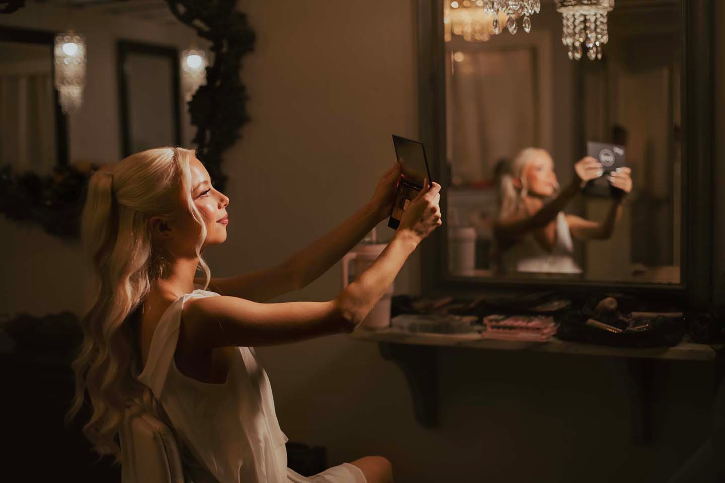 Blonde woman in white dress takes selfie with tablet in dimly lit vanity room with mirror reflection.