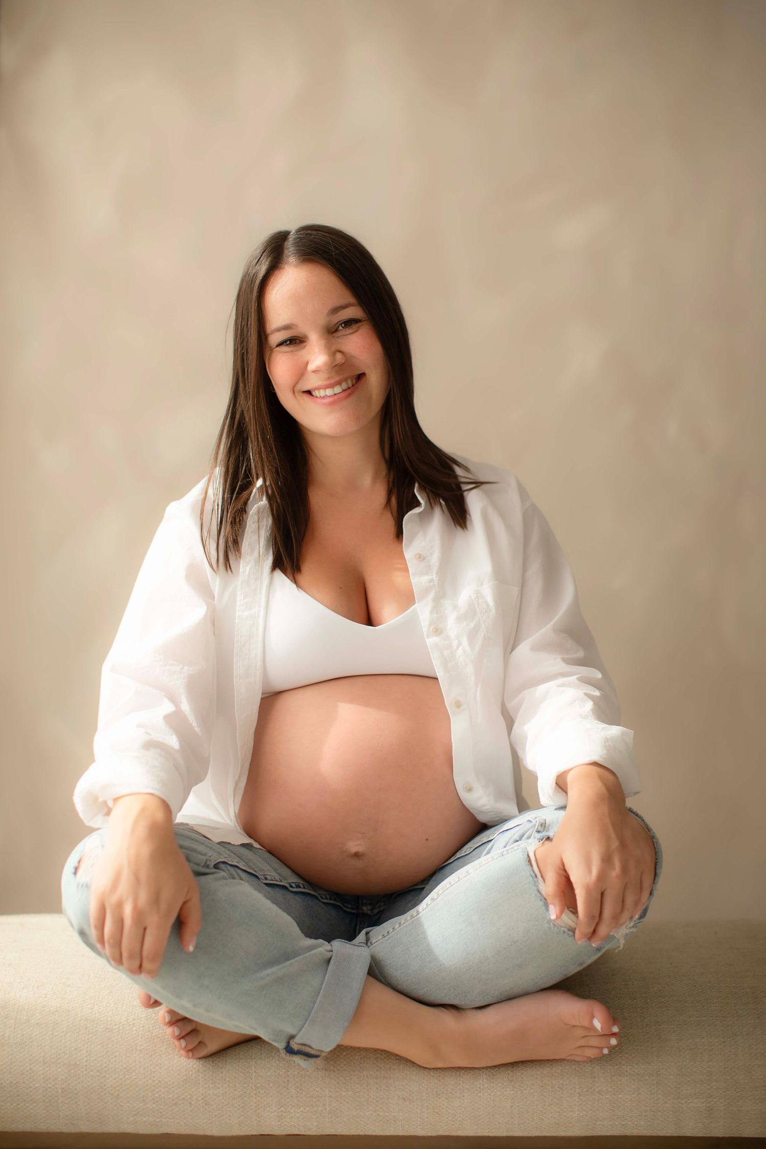 Smiling pregnant woman sitting cross-legged in white shirt and jeans, showing baby bump.