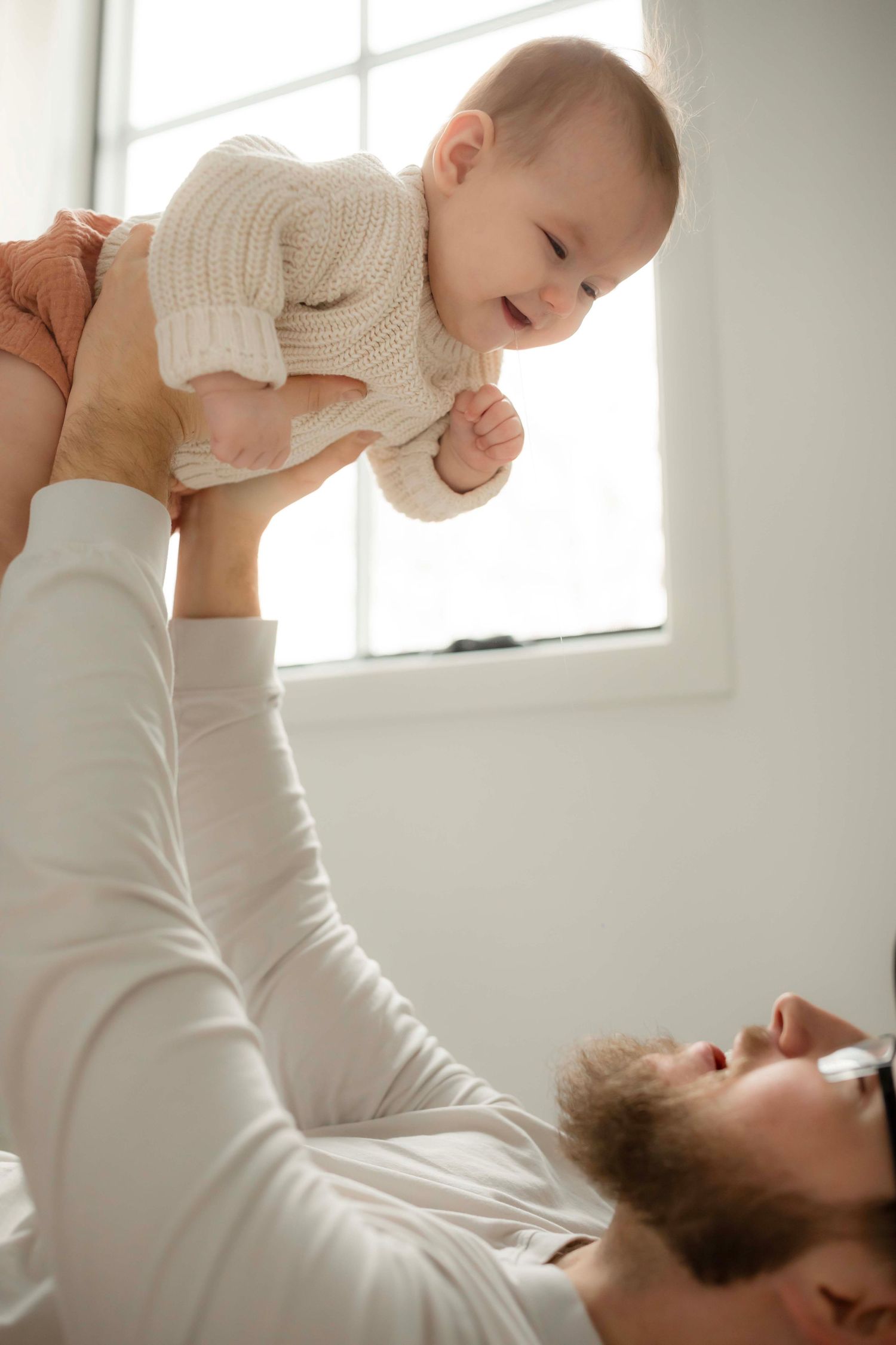 Parent lifts smiling baby overhead in playful sequence against bright window background.