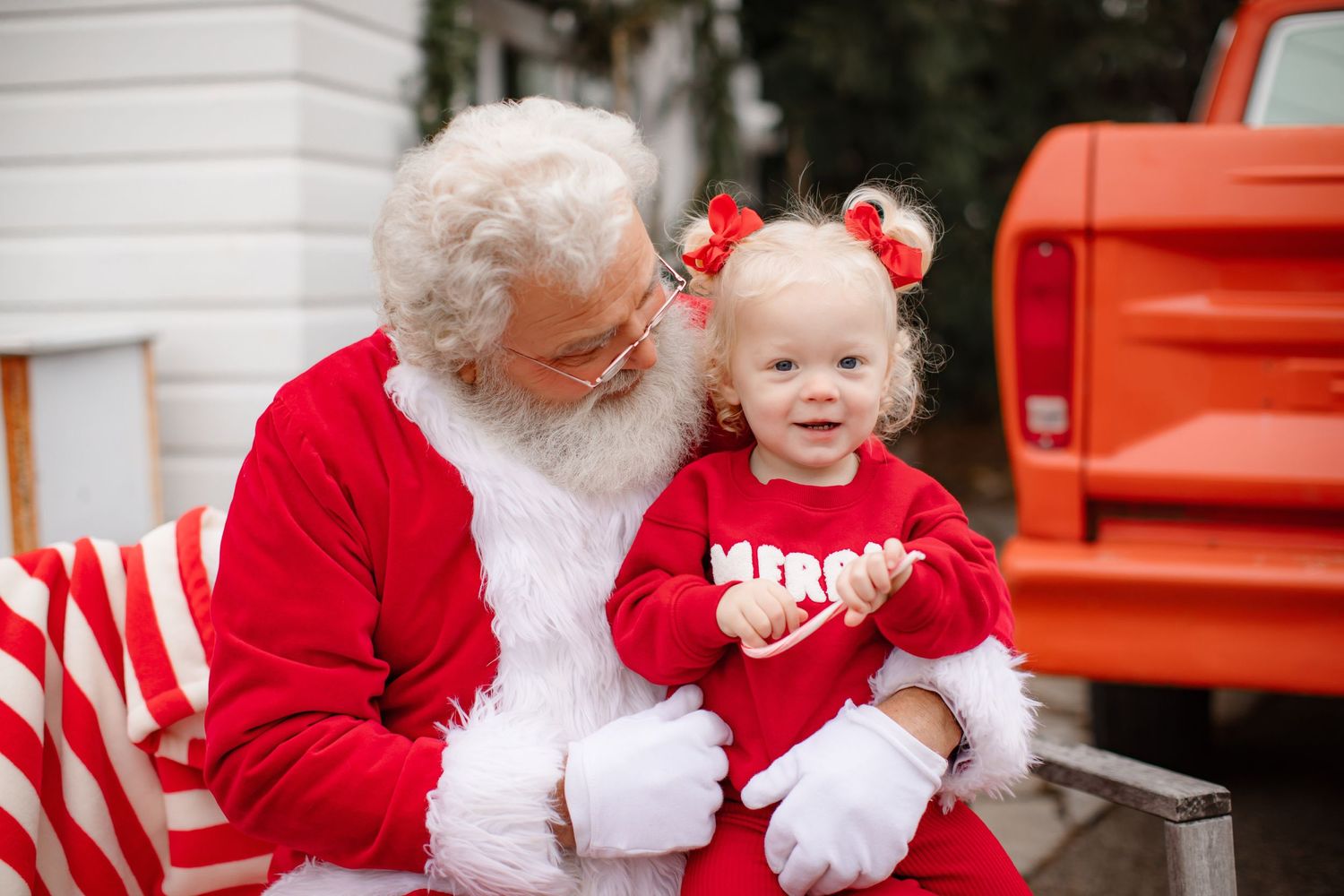 Santa and child in matching red outfits smile together near red truck and candy canes.