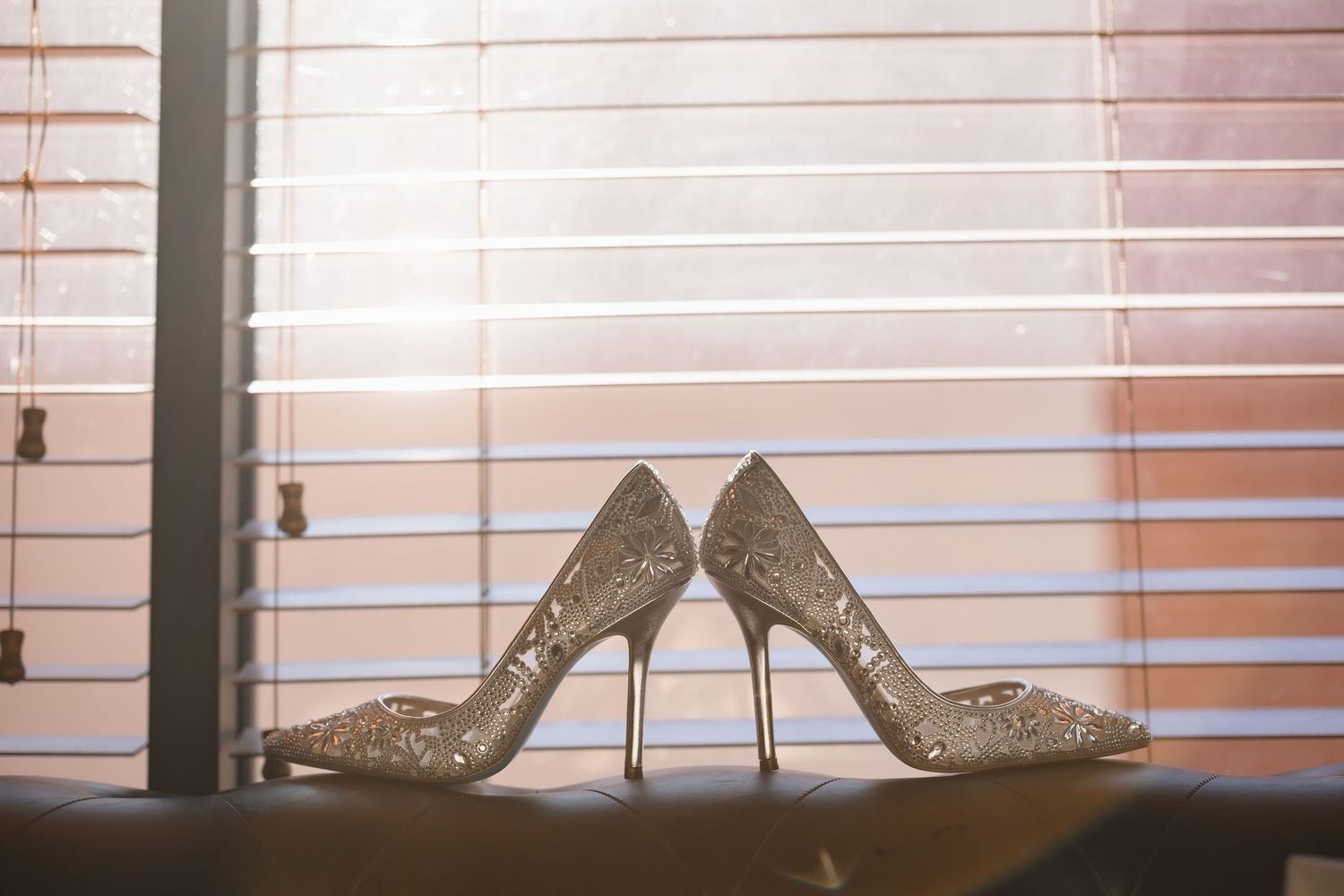 Sparkly silver wedding high heels positioned beside a window with venetian blinds.