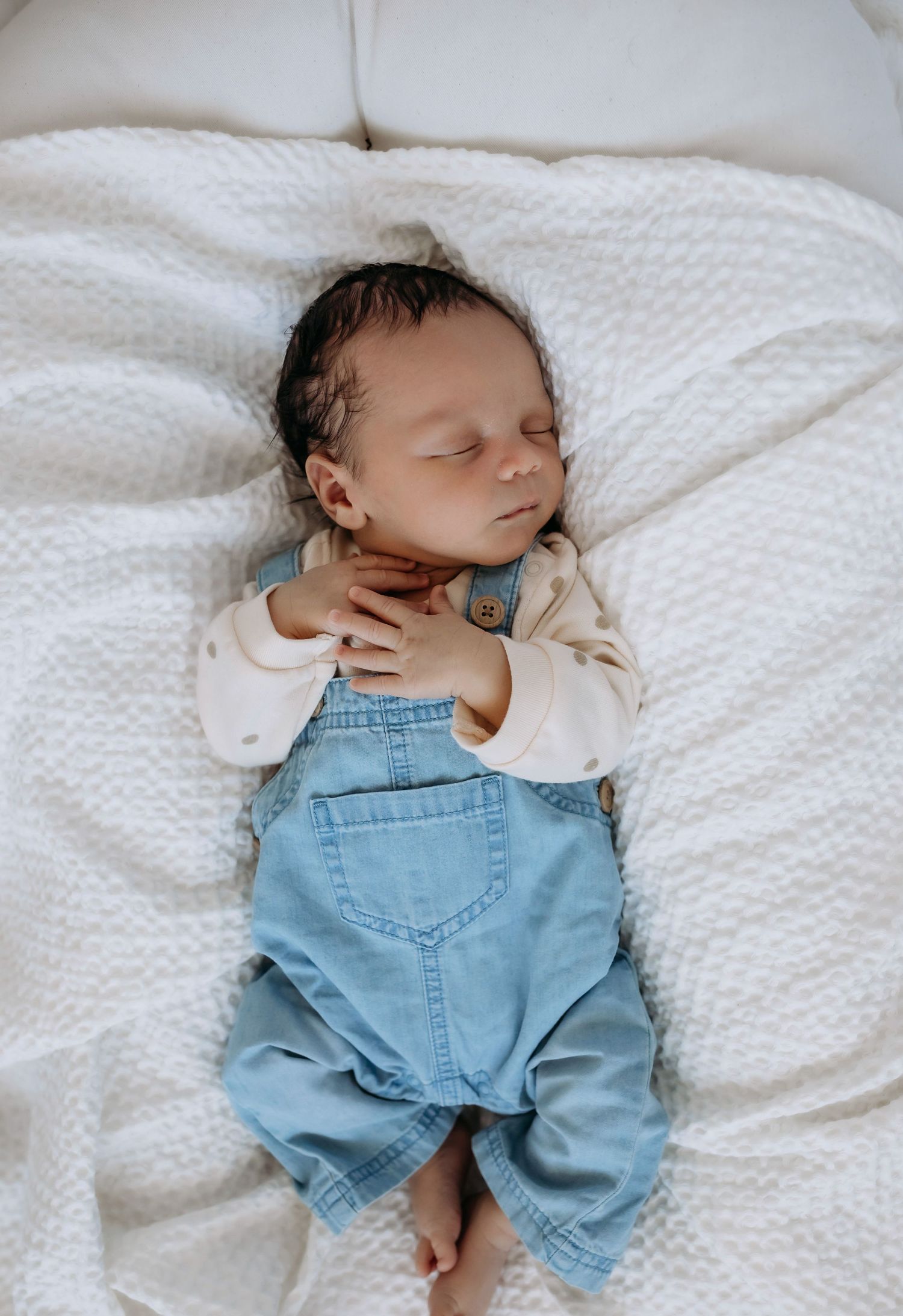 Sleeping newborn wearing denim overalls and white shirt on textured white blanket.