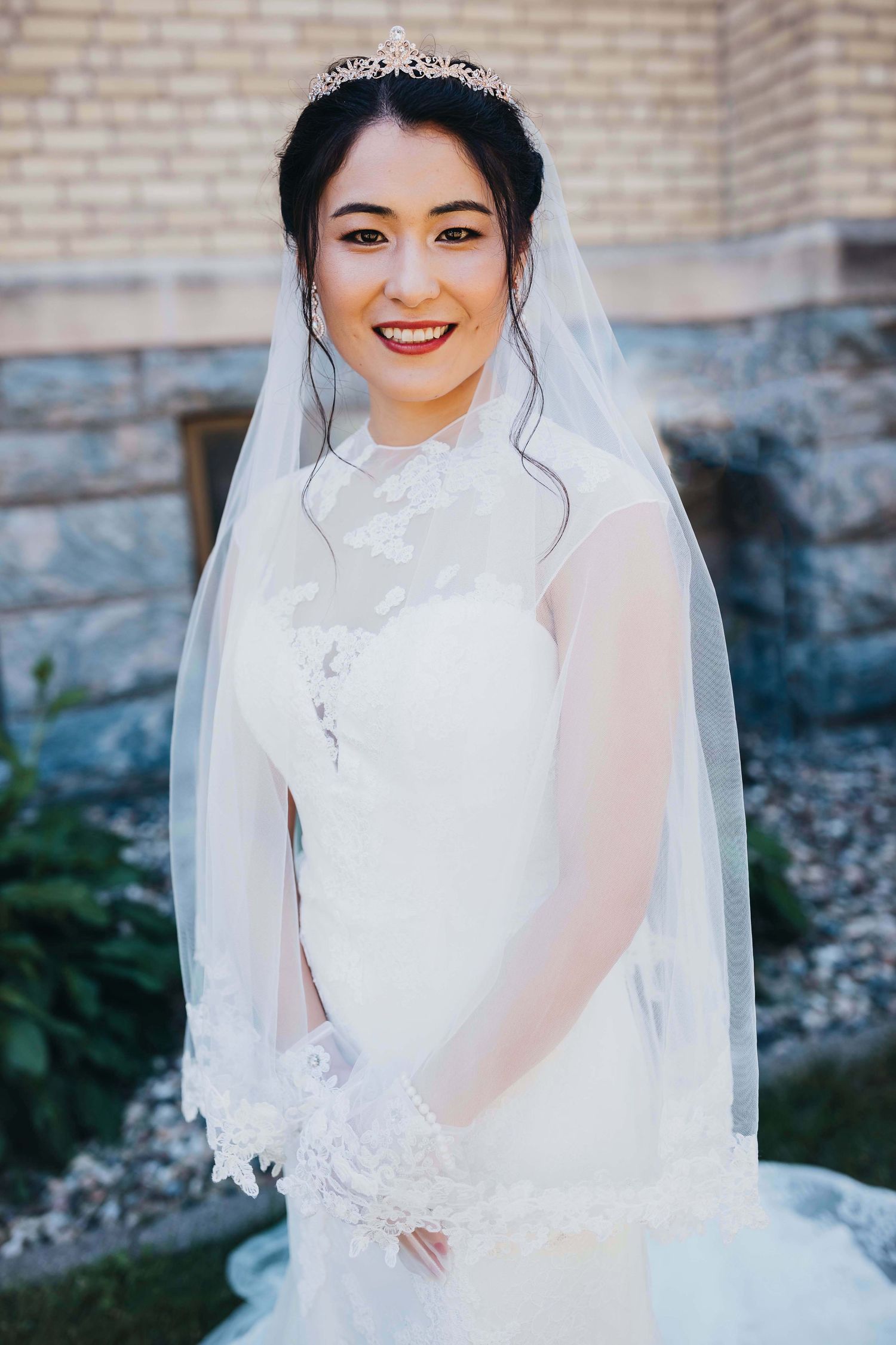 Elegant bridal portrait showcasing lace dress, veil and tiara against church wall.