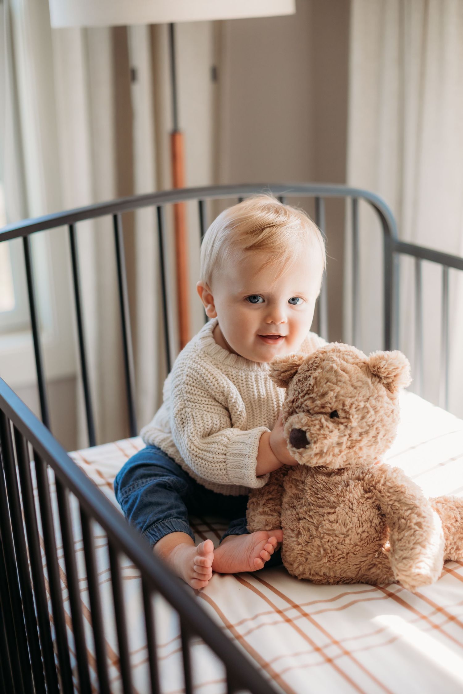 A baby in a cream sweater sits in a crib with a teddy bear.