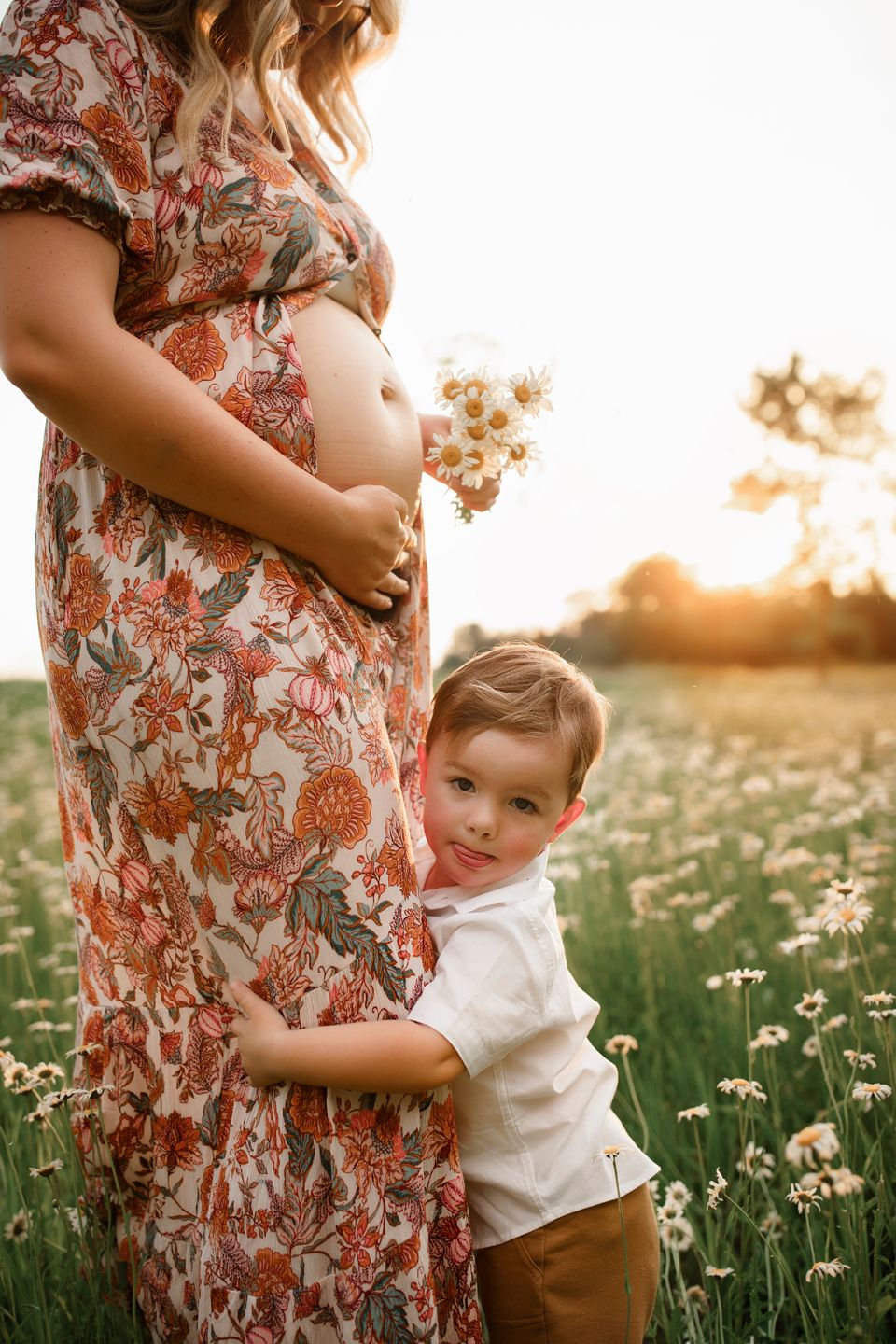A touching maternity scene shows a small child hugging a pregnant belly in a field of flowers at sunset.