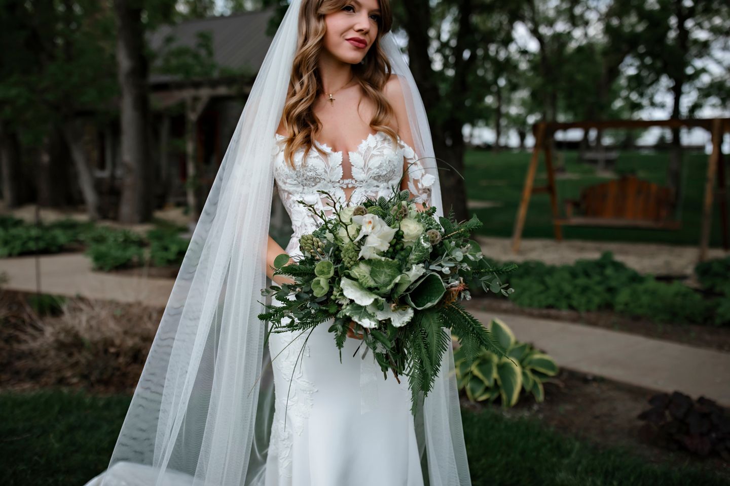 Bride in white lace gown holds lush greenery bouquet while standing outdoors with flowing veil.