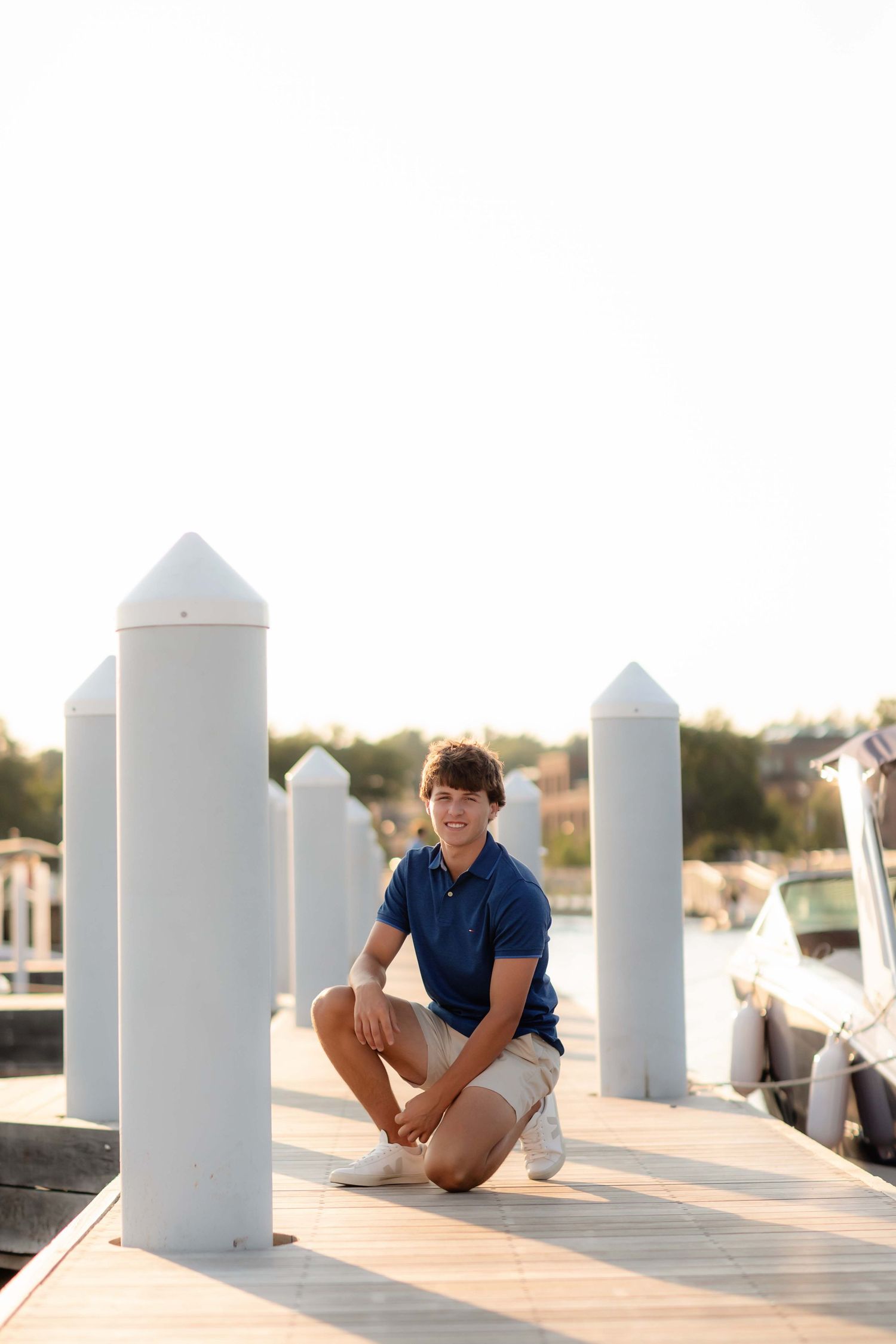 Teen boy in blue polo and khaki shorts crouching on a marina dock with white bollards at golden hour.