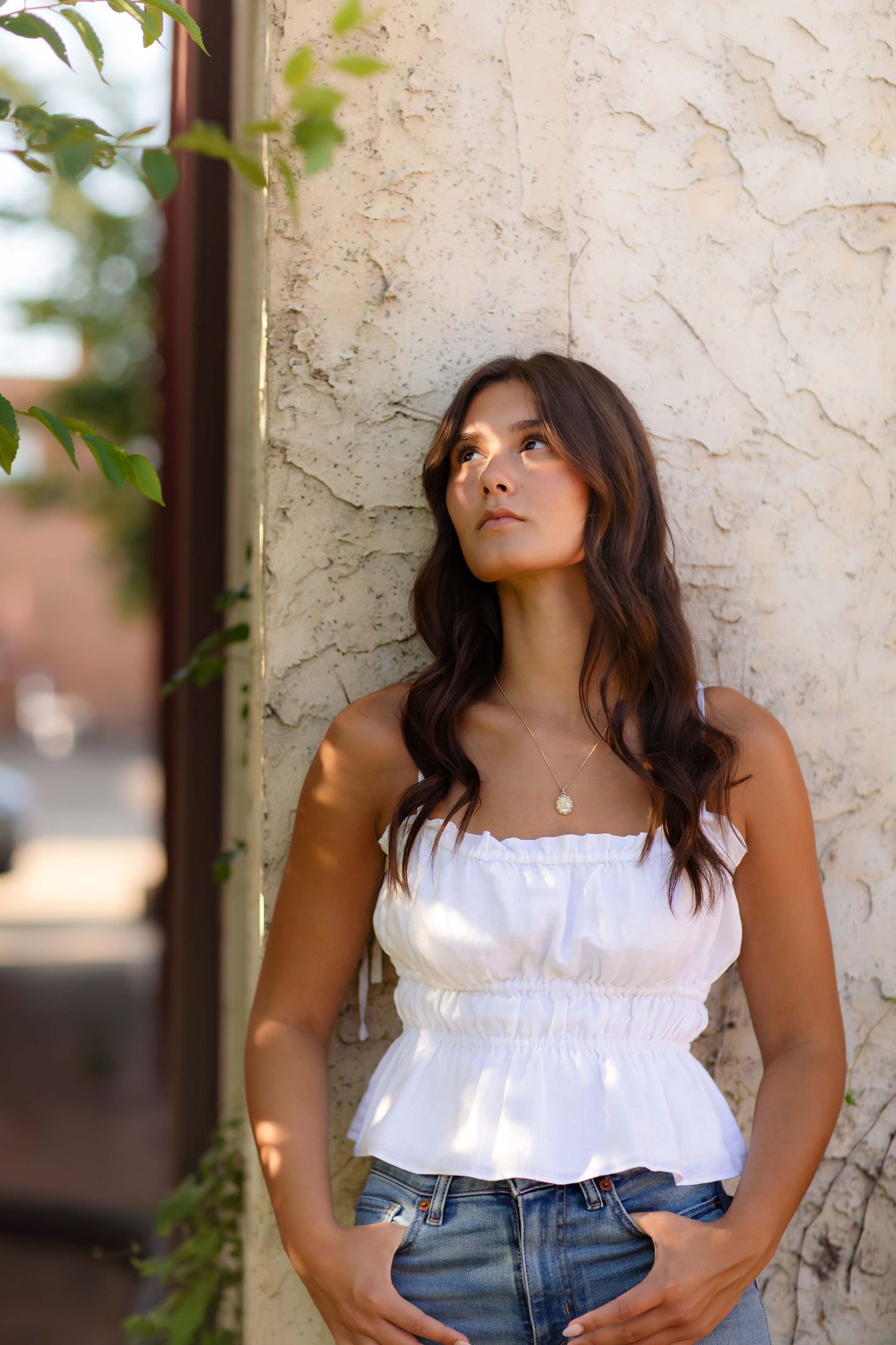 Young woman in white ruffle crop top and jeans leaning against textured wall outdoors, looking up thoughtfully.