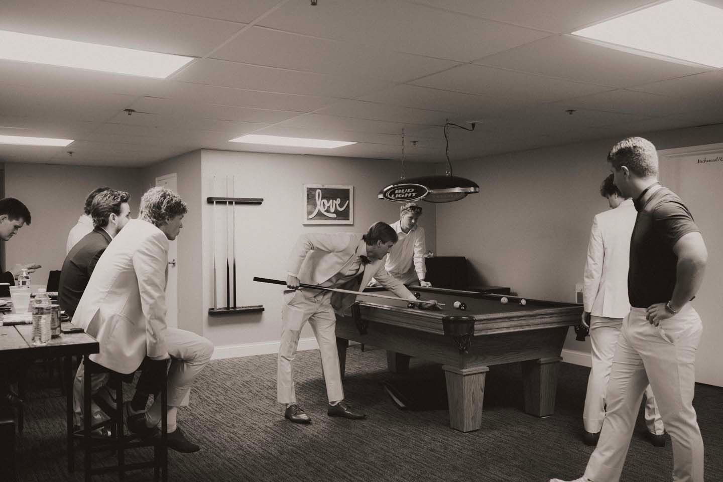 Groomsmen in white suits playing pool in a game room, captured in black and white photography.