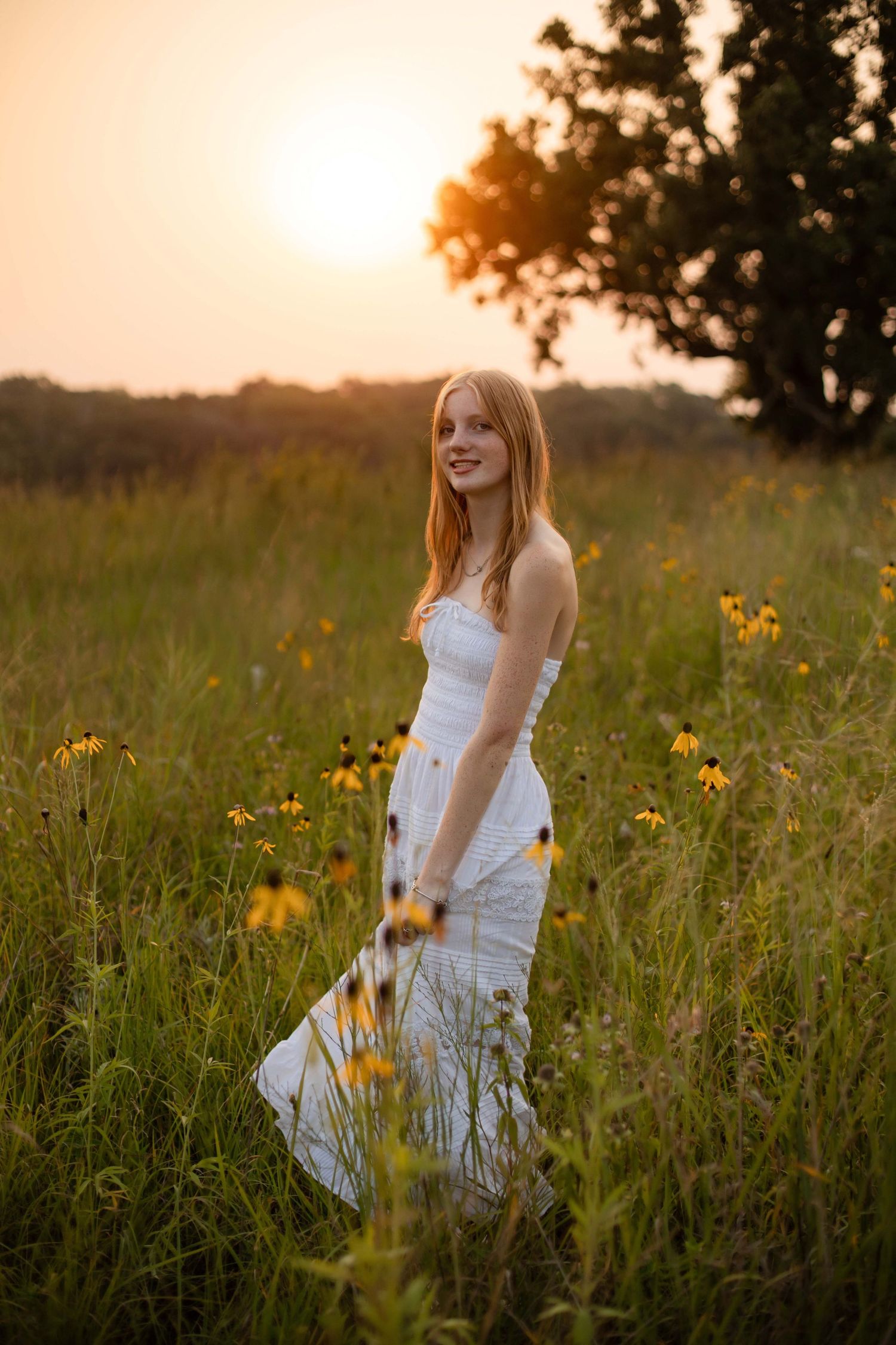 Young woman in white dress standing in wildflower field at golden sunset, smiling among yellow flowers.