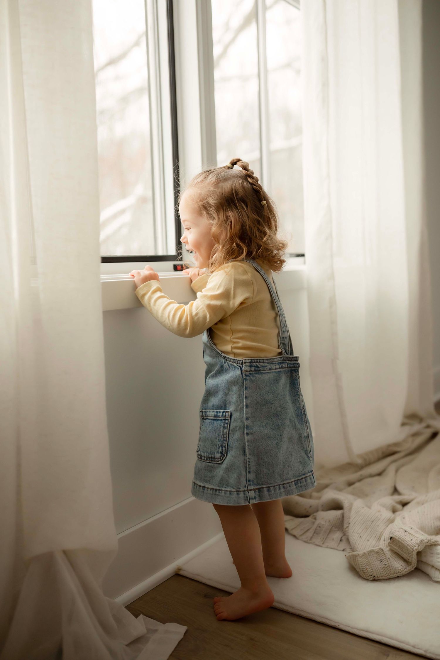 A toddler in denim overalls stands by a bright window with sunlight streaming through sheer curtains.