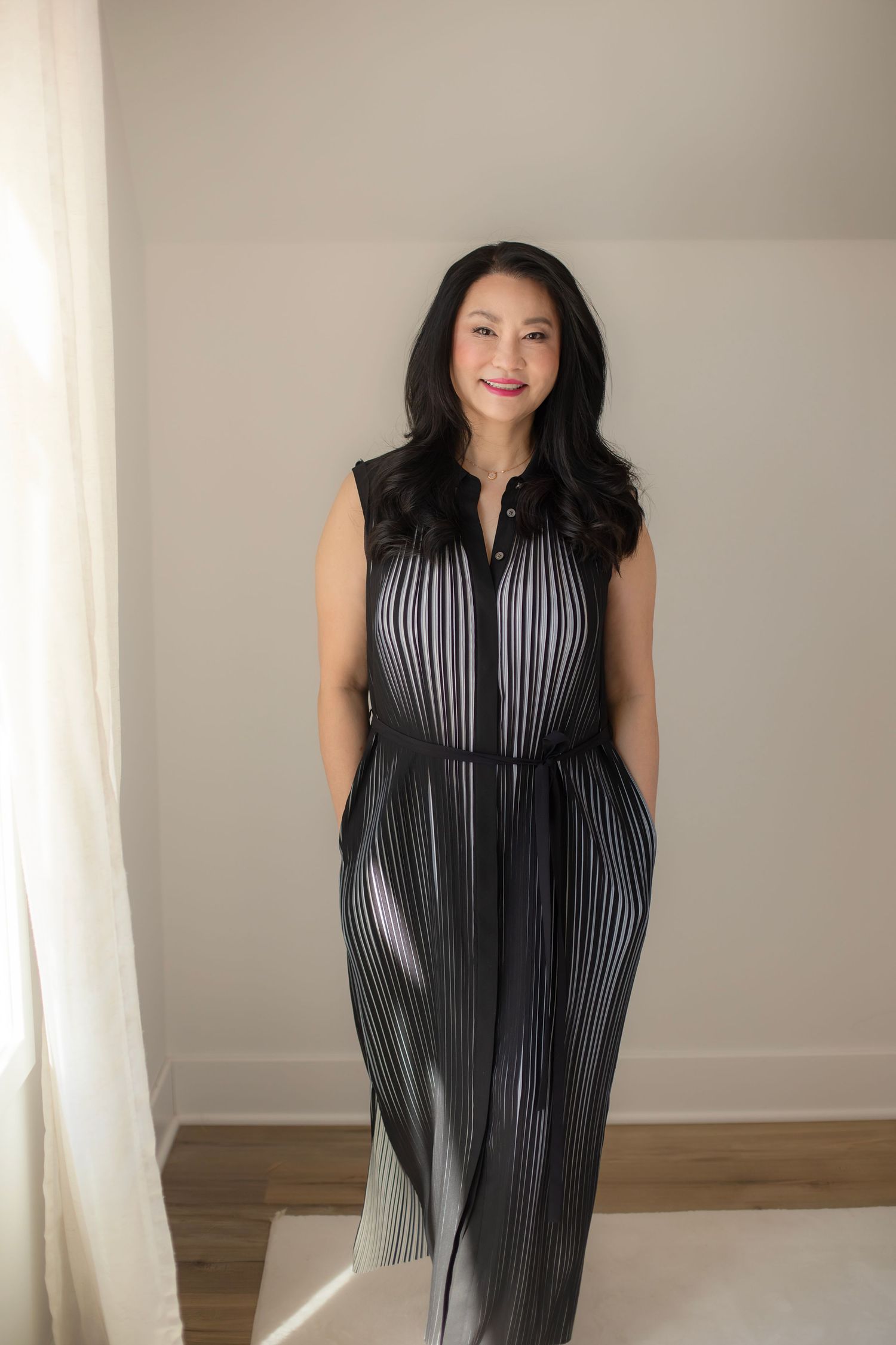 Elegant fashion poses in a black pleated dress with architectural details against a neutral wall.