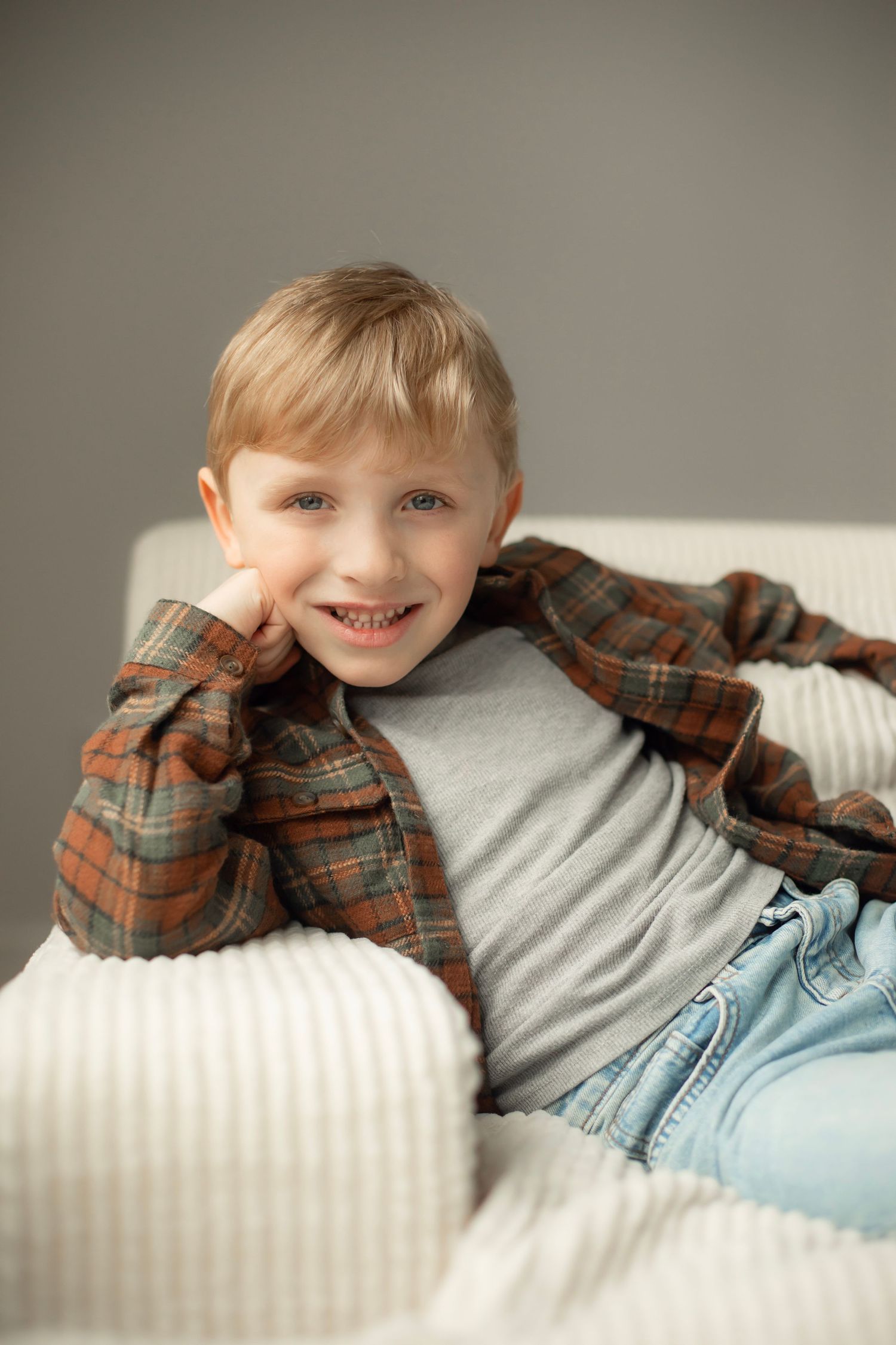 A child relaxes on white cushions wearing a plaid flannel shirt over a gray t-shirt and light denim jeans.