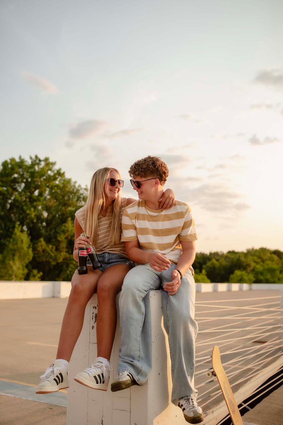 Young couple sitting together at a skate park during golden hour, sharing drinks and smiling.