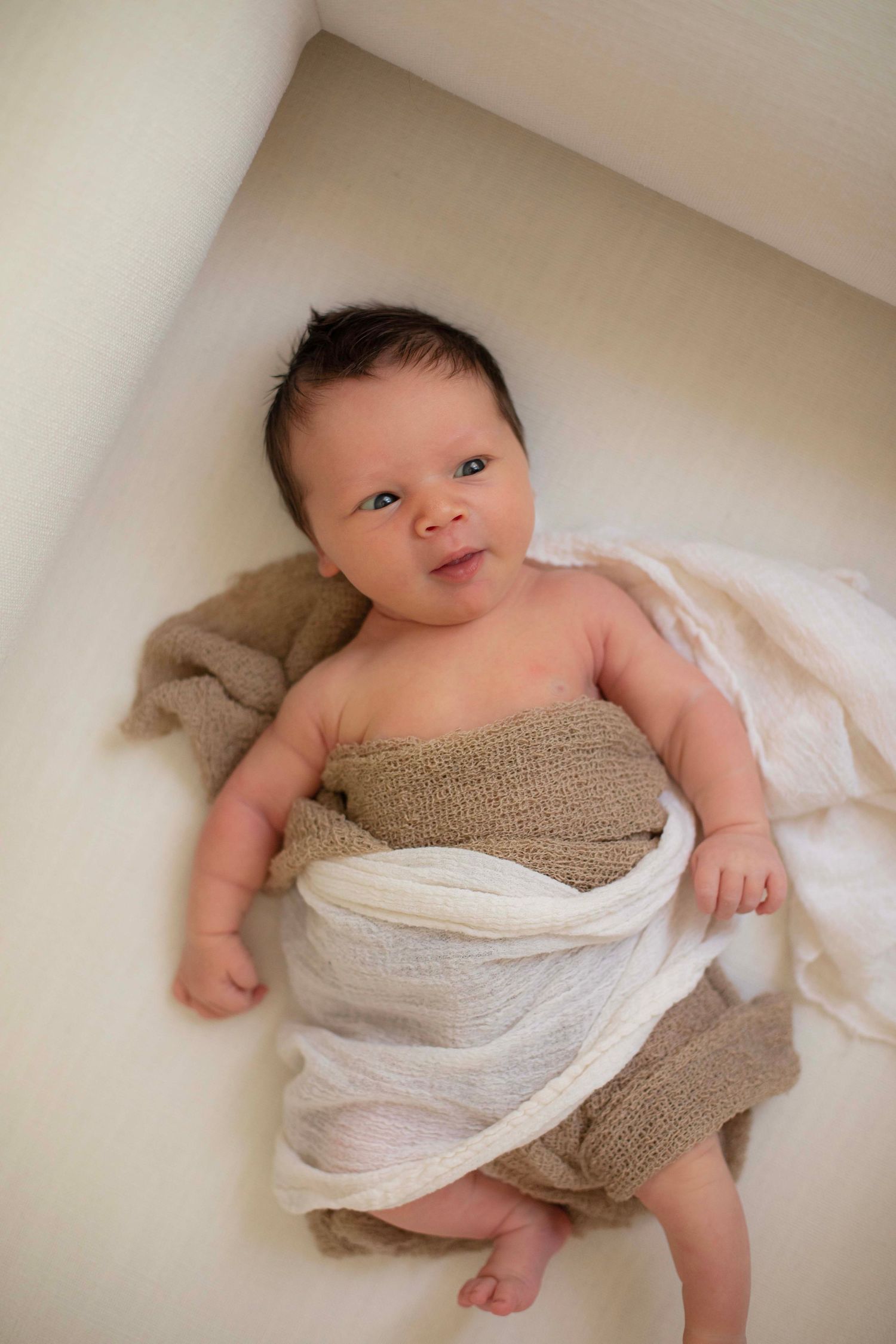 Newborn baby wrapped in beige and white knit blankets, lying on a white surface, looking up with dark hair.