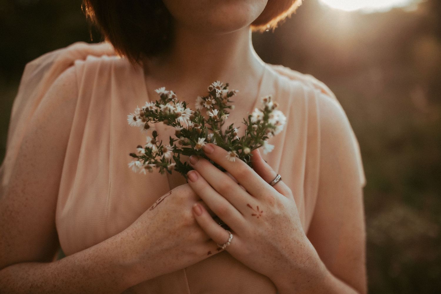 Hands gently holding small white wildflowers against a peachy-pink dress in golden light.