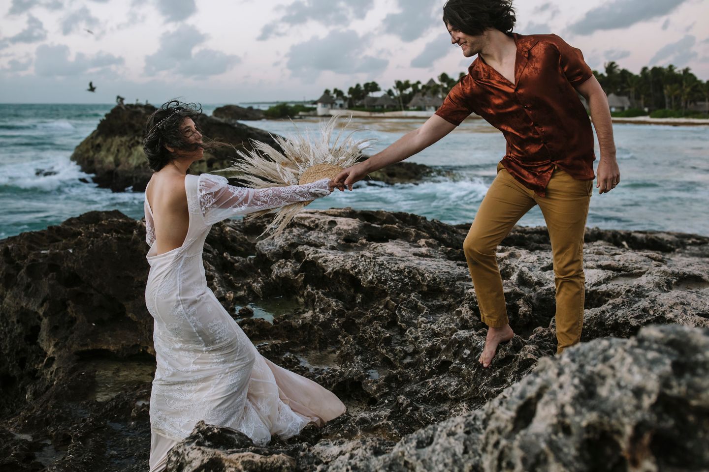 A couple in wedding attire helps each other across rocky coastal terrain with ocean waves in background.