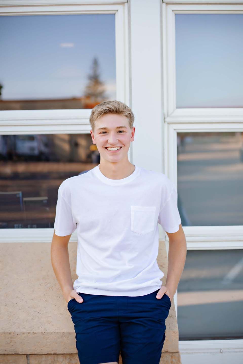 Young person in white t-shirt and navy shorts smiling against window backdrop on sunny day.